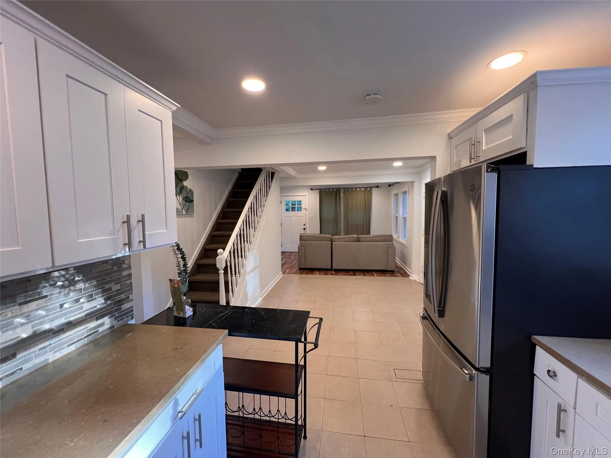 White shaker cabinetry with multi-tone tile backsplash, light toned countertop, stainless steel refrigerator, and tiled flooring White shaker cabinetry with multi-tone tile backsplash, light toned countertop, stainless steel refrigerator, and tiled flooring