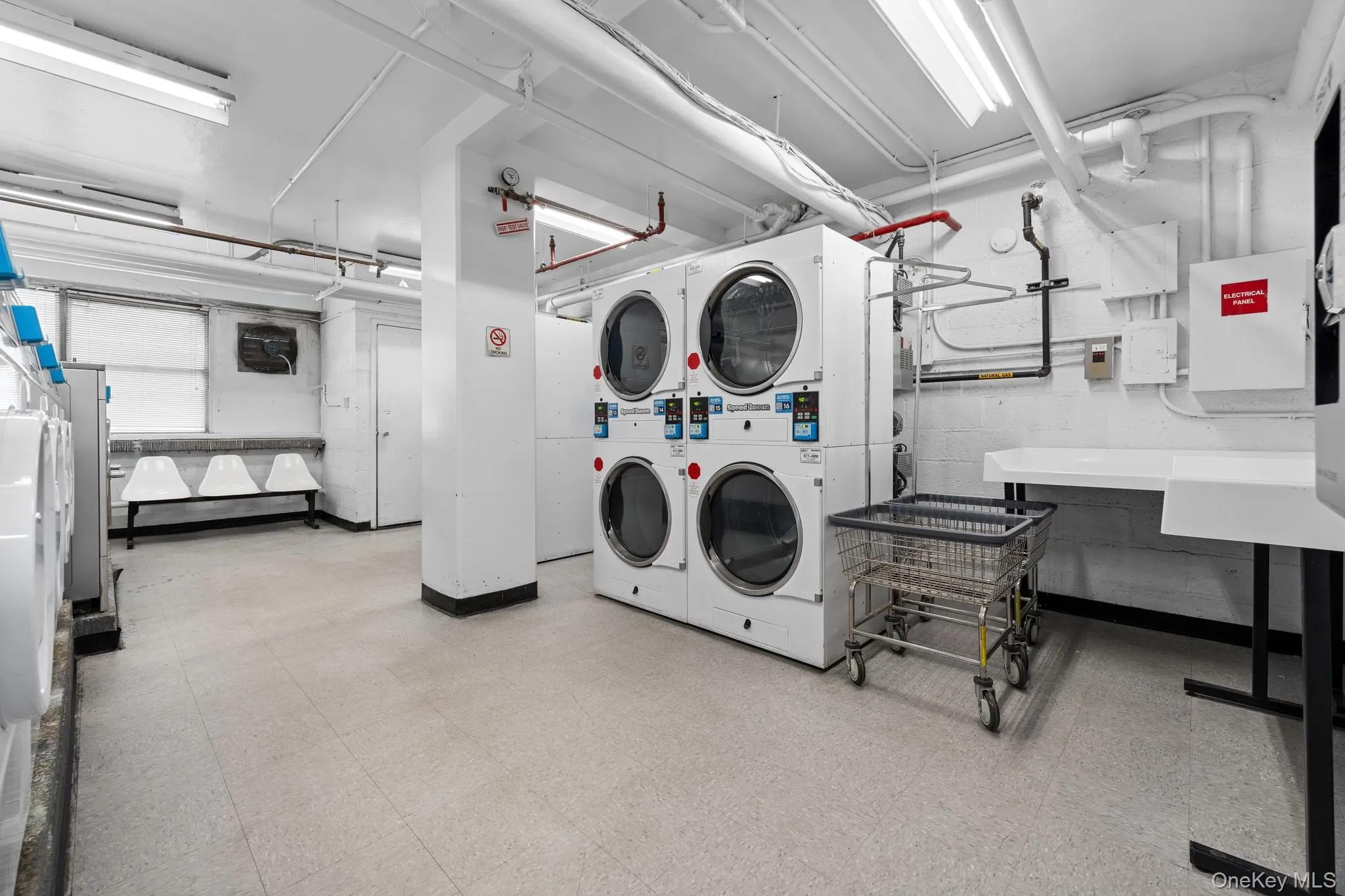 Communal laundry area with multiple large-capacity washing machines and dryers, featuring fluorescent lighting and a utility sink Communal laundry area with multiple large-capacity washing machines and dryers, featuring fluorescent lighting and a utility sink