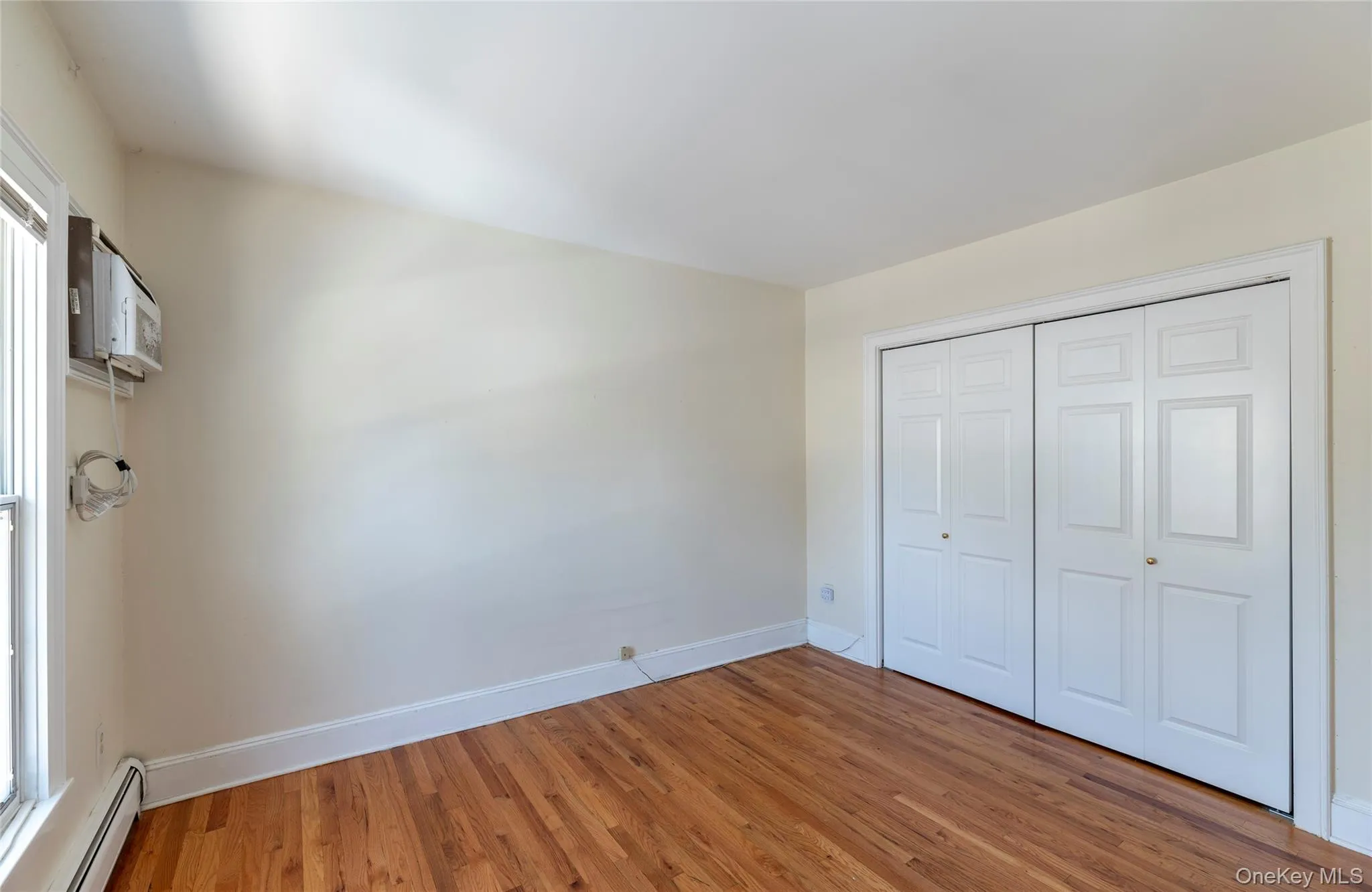 Unfurnished bedroom with a baseboard radiator, light wood finished floors, a closet, and a wall unit AC Unfurnished bedroom with a baseboard radiator, light wood finished floors, a closet, and a wall unit AC
