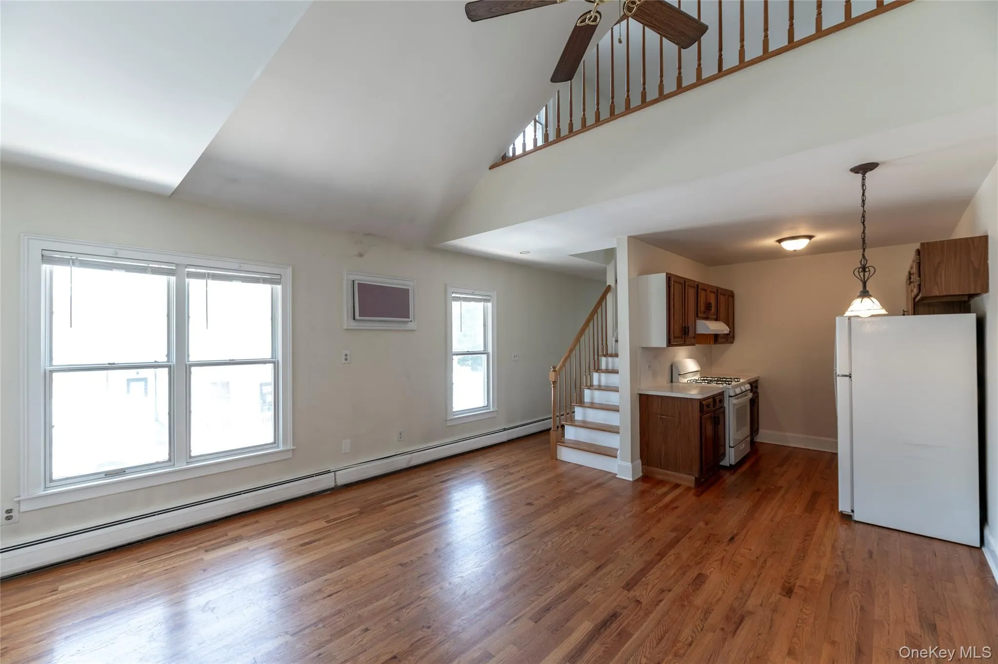 Unfurnished living room with ceiling fan, a baseboard radiator, wood finished floors, a towering ceiling, and stairs Unfurnished living room with ceiling fan, a baseboard radiator, wood finished floors, a towering ceiling, and stairs