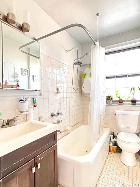White tiled walls featuring a built-in bathtub with a shower fixture, a wood-finish vanity with an integrated sink, a rectangular mirrored medicine cabinet, and a window providing natural illumination White tiled walls featuring a built-in bathtub with a shower fixture, a wood-finish vanity with an integrated sink, a rectangular mirrored medicine cabinet, and a window providing natural illumination