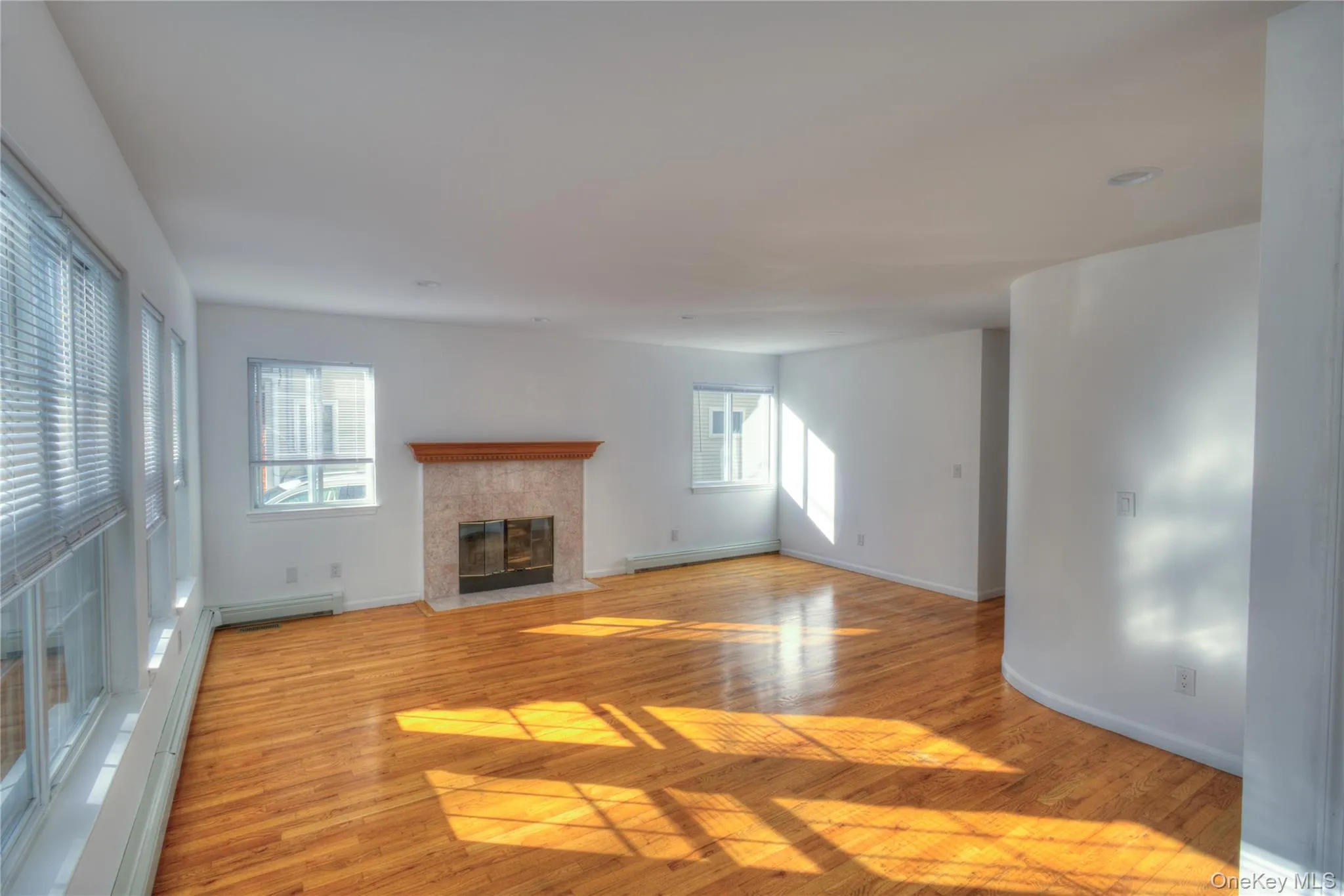 Hardwood flooring, multiple windows with blinds, built-in fireplace with stone surround and wood mantel, white walls, and recessed lighting Hardwood flooring, multiple windows with blinds, built-in fireplace with stone surround and wood mantel, white walls, and recessed lighting
