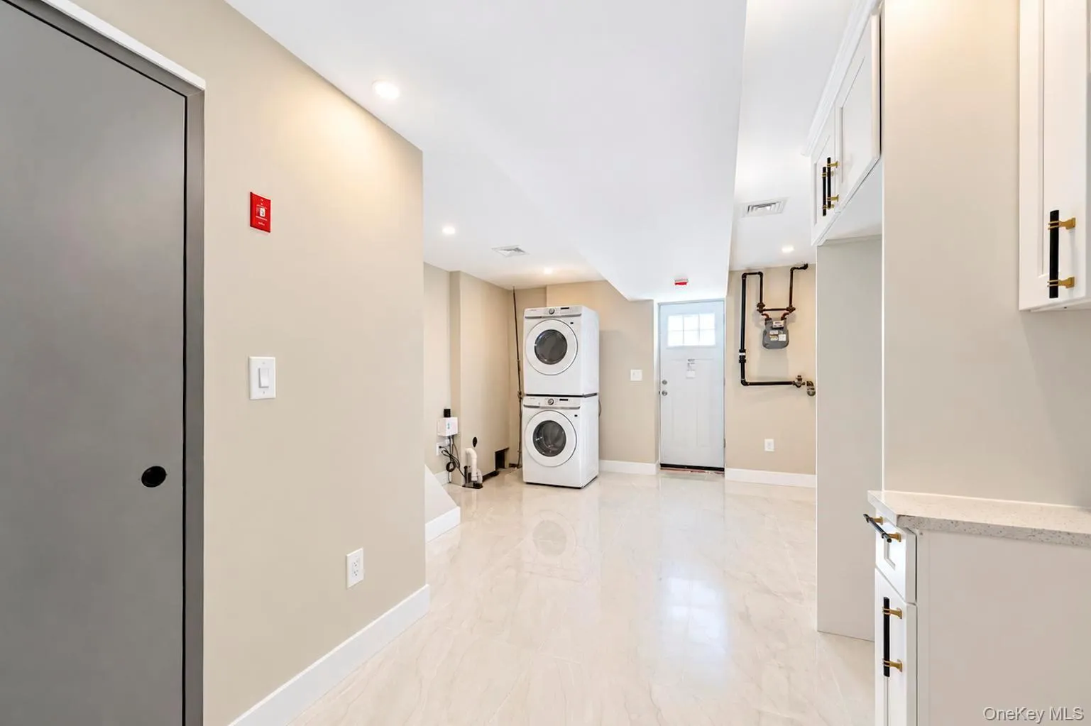 White tile flooring, white shaker cabinetry with black and gold hardware, recessed lighting, and a stackable washer and dryer unit. White tile flooring, white shaker cabinetry with black and gold hardware, recessed lighting, and a stackable washer and dryer unit.