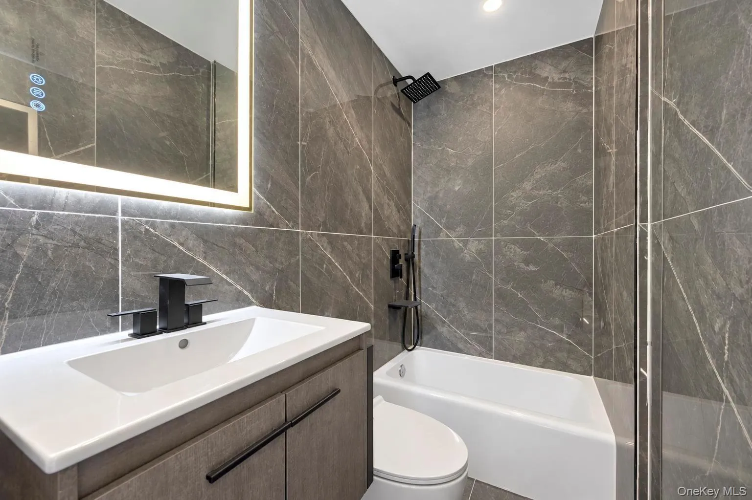 3rd Bathroom featuring floor-to-ceiling gray veined tile, an illuminated vanity mirror, a single-sink vanity with a flat-panel cabinet, a white bathtub, and a glass shower partition 3rd Bathroom featuring floor-to-ceiling gray veined tile, an illuminated vanity mirror, a single-sink vanity with a flat-panel cabinet, a white bathtub, and a glass shower partition