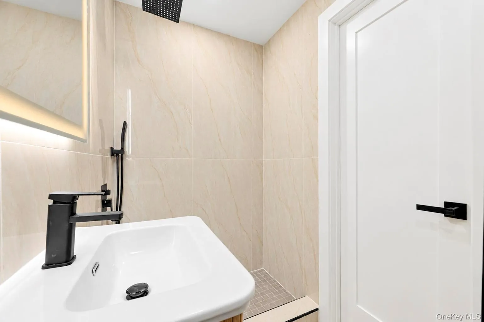 Wall-mounted, vanity sink with matte black faucet, illuminated mirror, and tiled shower surround with rainfall showerhead Wall-mounted, vanity sink with matte black faucet, illuminated mirror, and tiled shower surround with rainfall showerhead
