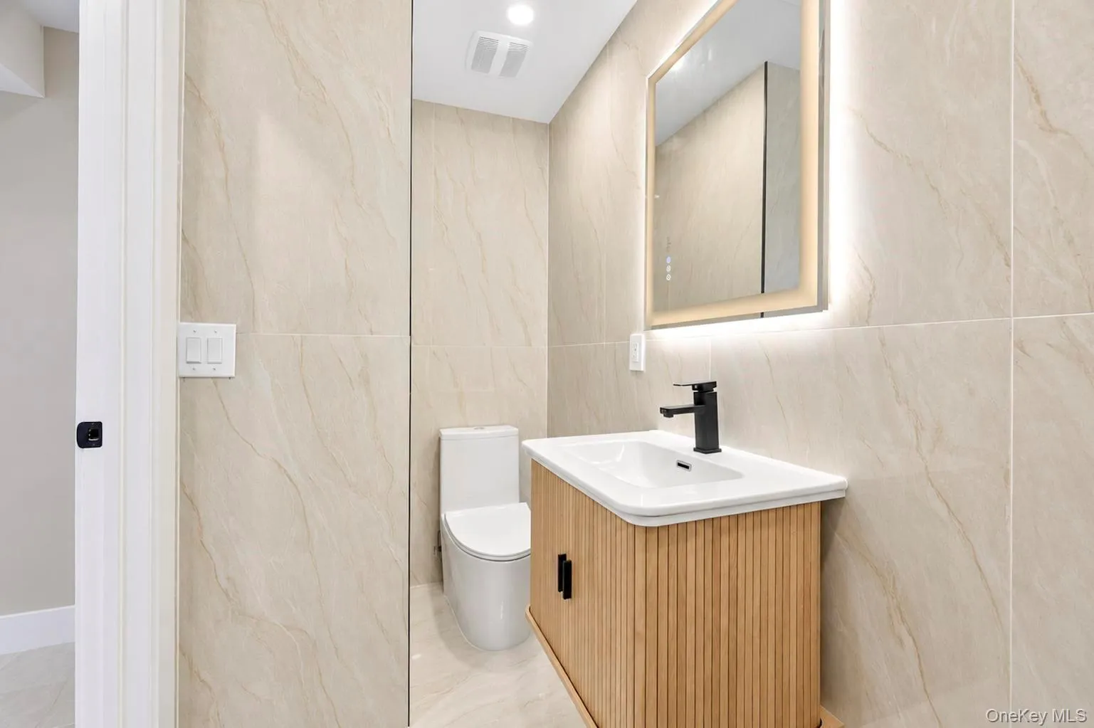 Bathroom, with full-height tiled walls, a wood-finish vanity with fluted detailing, a white integrated sink, a matte black faucet, and an illuminated mirrored medicine cabinet Bathroom, with full-height tiled walls, a wood-finish vanity with fluted detailing, a white integrated sink, a matte black faucet, and an illuminated mirrored medicine cabinet