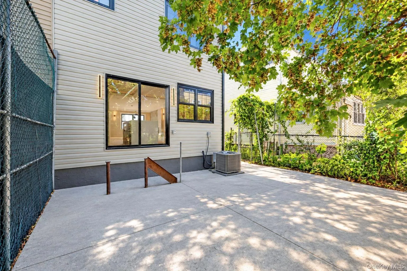 Poured concrete patio with lush perimeter landscaping, chain-link fencing, and exterior wall sconces Poured concrete patio with lush perimeter landscaping, chain-link fencing, and exterior wall sconces
