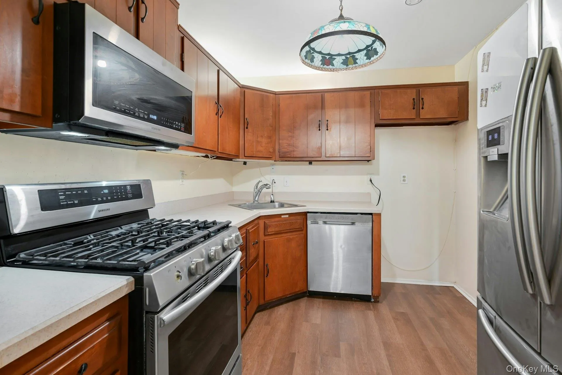 Kitchen with appliances with stainless steel finishes, light wood finished floors, light countertops, brown cabinetry, and hanging light fixtures Kitchen with appliances with stainless steel finishes, light wood finished floors, light countertops, brown cabinetry, and hanging light fixtures