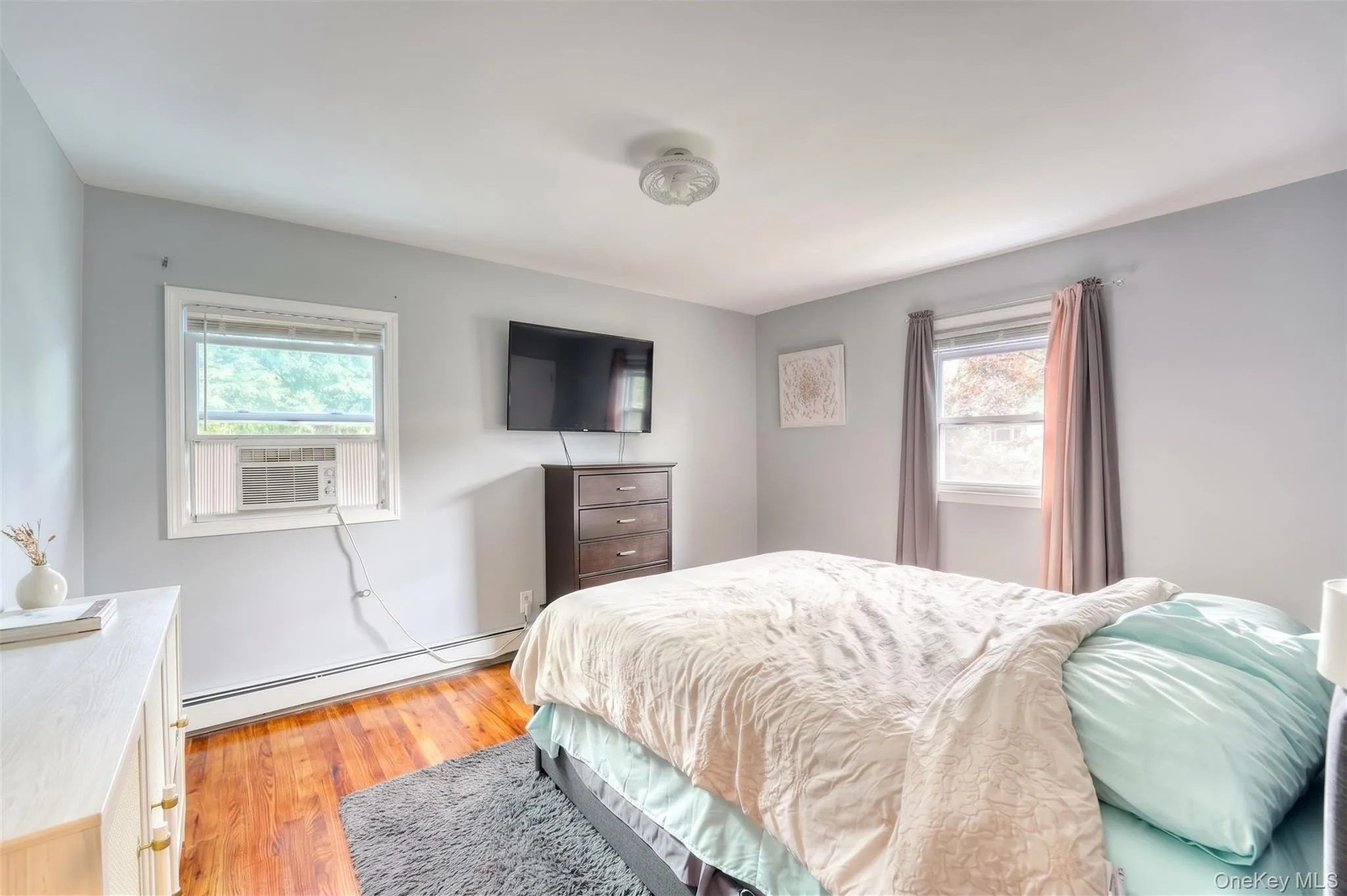 Bright and comfortable bedroom featuring hardwood floors, soft gray walls, and large windows that fill the space with natural light. The room includes a mounted flat-screen TV, dark wood dresser, and a cozy bed dressed in neutral tones with mint accents. Thoughtful touches like coordinating curtains, framed artwork, and a plush area rug create a relaxing, modern retreat perfect for everyday living. Bright and comfortable bedroom featuring hardwood floors, soft gray walls, and large windows that fill the space with natural light. The room includes a mounted flat-screen TV, dark wood dresser, and a cozy bed dressed in neutral tones with mint accents. Thoughtful touches like coordinating curtains, framed artwork, and a plush area rug create a relaxing, modern retreat perfect for everyday living.