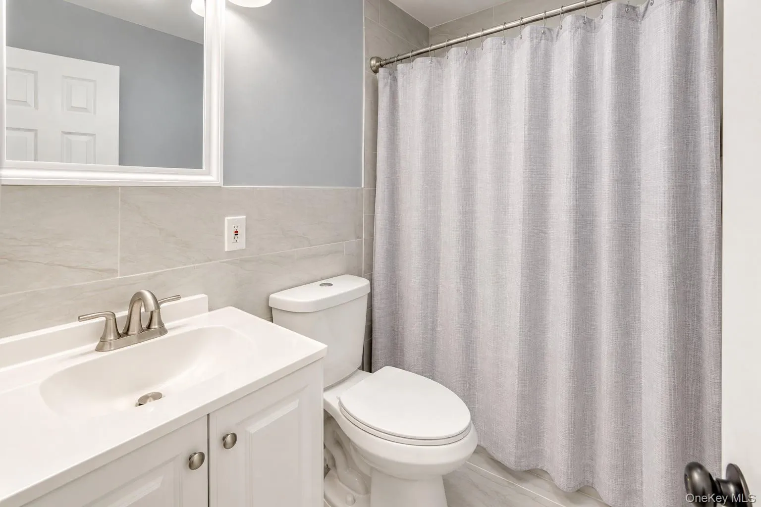 Modern full bath featuring a white vanity with brushed-nickel fixtures, matching hardware, and a sleek integrated sink. Light gray tile walls and flooring complement the soft gray paint for a fresh, neutral look. Includes a standard tub with a textured gray shower curtain, updated lighting, and a framed mirror for a bright, polished finish. Modern full bath featuring a white vanity with brushed-nickel fixtures, matching hardware, and a sleek integrated sink. Light gray tile walls and flooring complement the soft gray paint for a fresh, neutral look. Includes a standard tub with a textured gray shower curtain, updated lighting, and a framed mirror for a bright, polished finish.