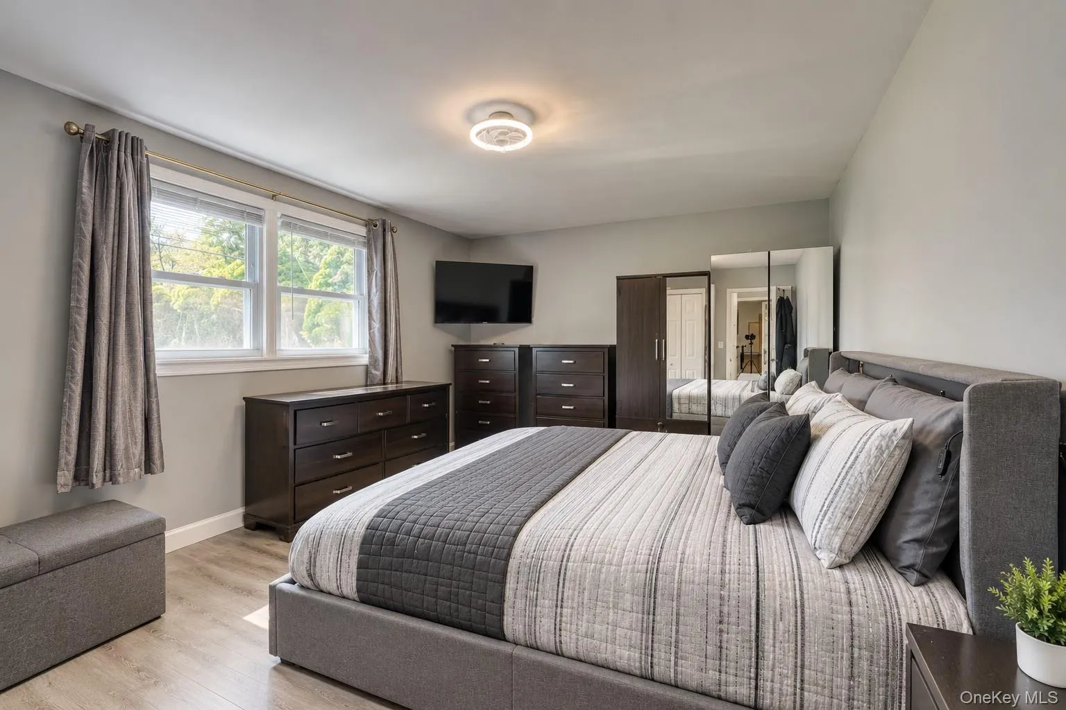 Elegant and spacious bedroom showcasing a modern design with clean lines and neutral tones. The room features a large upholstered bed with coordinated bedding, dark wood furniture, and mirrored wardrobe doors that enhance light and depth. Wide windows with gray curtains fill the space with natural light, while the sleek ceiling fixture adds a contemporary touch. This inviting retreat blends comfort, style, and functionality — perfect for relaxing or unwinding after a long day. Elegant and spacious bedroom showcasing a modern design with clean lines and neutral tones. The room features a large upholstered bed with coordinated bedding, dark wood furniture, and mirrored wardrobe doors that enhance light and depth. Wide windows with gray curtains fill the space with natural light, while the sleek ceiling fixture adds a contemporary touch. This inviting retreat blends comfort, style, and functionality — perfect for relaxing or unwinding after a long day.