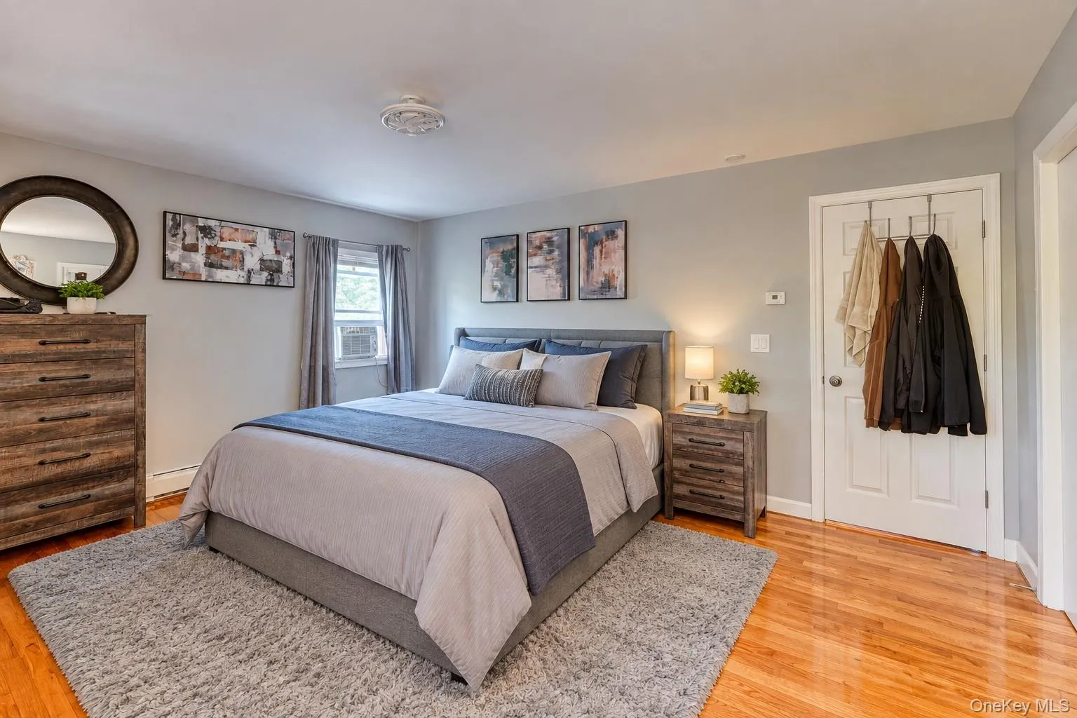 Inviting primary bedroom featuring warm hardwood floors, soft gray walls, and abundant natural light. The space includes a plush area rug, modern wood-grain furniture, and a stylish upholstered bed with layered bedding in calming neutral tones. Thoughtful details like coordinated artwork, brushed-nickel accents, and contemporary lighting create a relaxing, move-in-ready retreat that blends comfort and sophistication. Inviting primary bedroom featuring warm hardwood floors, soft gray walls, and abundant natural light. The space includes a plush area rug, modern wood-grain furniture, and a stylish upholstered bed with layered bedding in calming neutral tones. Thoughtful details like coordinated artwork, brushed-nickel accents, and contemporary lighting create a relaxing, move-in-ready retreat that blends comfort and sophistication.