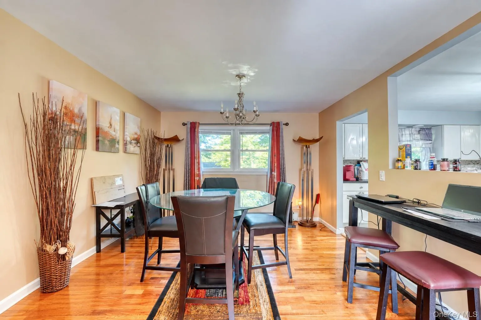 Warm and inviting dining area featuring hardwood floors, neutral walls, and a modern glass-top table with four leather chairs. Natural light pours in through double windows framed by red and beige curtains, highlighting elegant décor elements like abstract wall art, tall wicker vases, and sculptural floor lamps. A chandelier adds a touch of sophistication, while the open layout connects seamlessly to the kitchen for easy entertaining. Warm and inviting dining area featuring hardwood floors, neutral walls, and a modern glass-top table with four leather chairs. Natural light pours in through double windows framed by red and beige curtains, highlighting elegant décor elements like abstract wall art, tall wicker vases, and sculptural floor lamps. A chandelier adds a touch of sophistication, while the open layout connects seamlessly to the kitchen for easy entertaining.