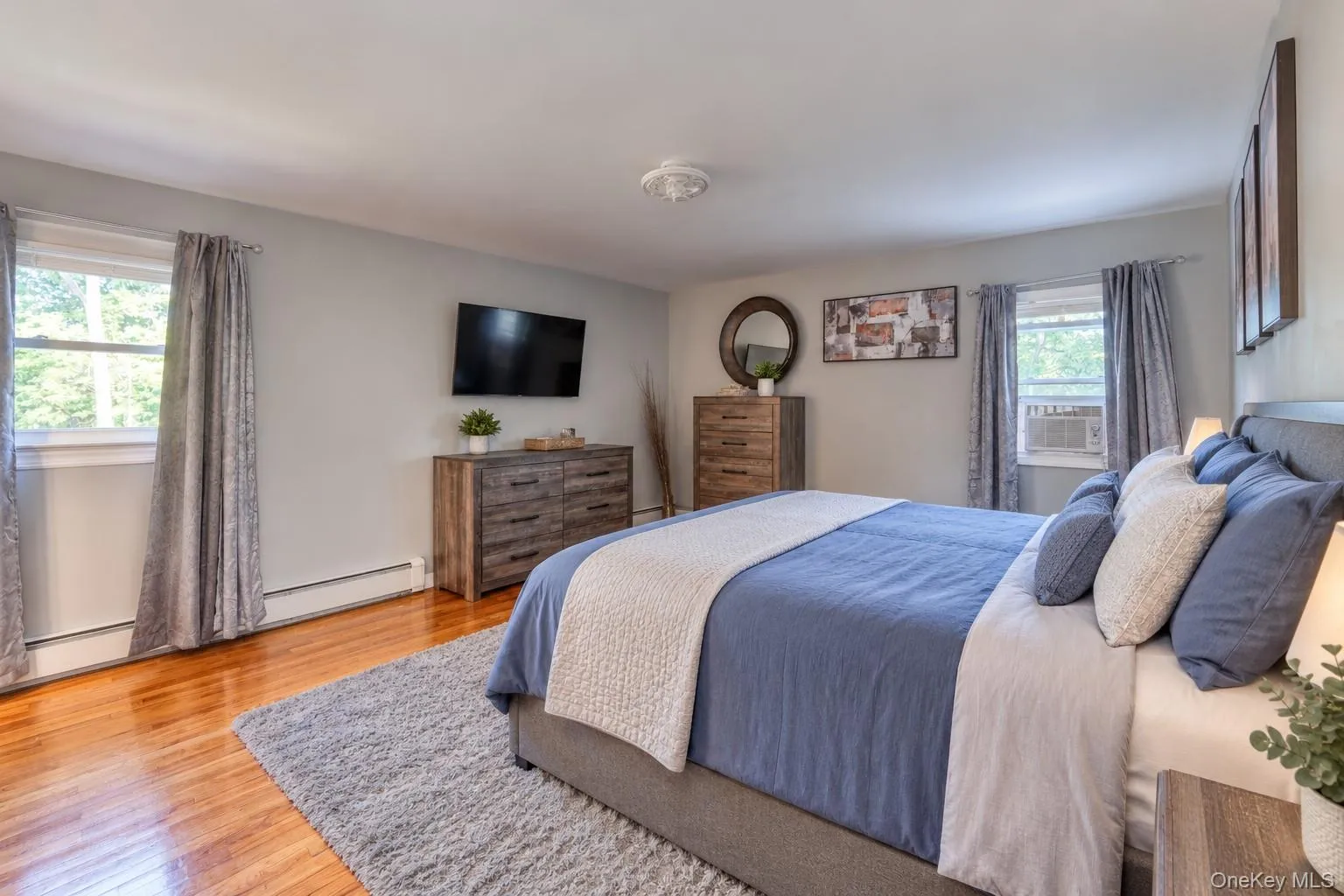 Spacious primary bedroom with en-suite bath, featuring warm hardwood floors and soft gray walls for a serene atmosphere. The room includes a large upholstered bed dressed in blue and white linens, complemented by matching wood-grain furniture and modern décor. Dual windows bring in natural light, while a mounted flat-screen TV and stylish accents create a comfortable, contemporary retreat ideal for relaxation. Spacious primary bedroom with en-suite bath, featuring warm hardwood floors and soft gray walls for a serene atmosphere. The room includes a large upholstered bed dressed in blue and white linens, complemented by matching wood-grain furniture and modern décor. Dual windows bring in natural light, while a mounted flat-screen TV and stylish accents create a comfortable, contemporary retreat ideal for relaxation.