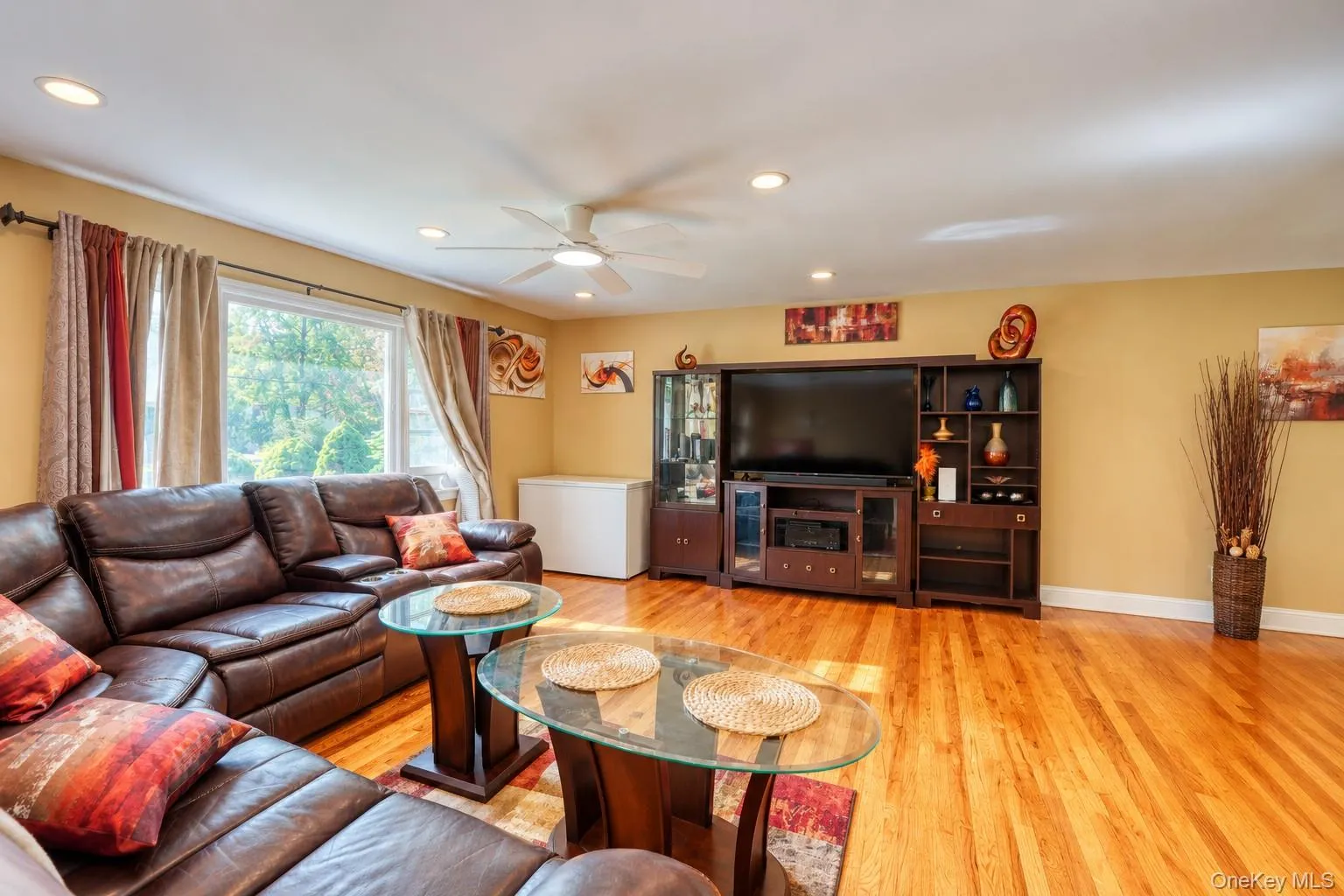 Inviting living room featuring rich hardwood floors, warm neutral walls, and a large picture window that fills the space with natural light. The room is anchored by a dark leather sectional sofa accented with vibrant red and orange pillows, complemented by glass-top coffee tables and a modern entertainment center with built-in shelving. Recessed lighting and a ceiling fan enhance comfort and brightness, while tasteful artwork and decorative touches create a cozy, stylish atmosphere perfect for relaxing or entertaining. Inviting living room featuring rich hardwood floors, warm neutral walls, and a large picture window that fills the space with natural light. The room is anchored by a dark leather sectional sofa accented with vibrant red and orange pillows, complemented by glass-top coffee tables and a modern entertainment center with built-in shelving. Recessed lighting and a ceiling fan enhance comfort and brightness, while tasteful artwork and decorative touches create a cozy, stylish atmosphere perfect for relaxing or entertaining.