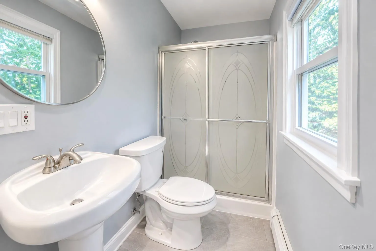 Bright, modern bathroom featuring a pedestal sink with brushed-nickel fixtures, a round mirror, and a frosted glass shower enclosure with decorative etching. Light gray walls and tile flooring create a clean, contemporary look, complemented by double windows that fill the space with natural light. This updated bath blends style and function, offering a fresh, airy feel ideal for today’s buyers. Bright, modern bathroom featuring a pedestal sink with brushed-nickel fixtures, a round mirror, and a frosted glass shower enclosure with decorative etching. Light gray walls and tile flooring create a clean, contemporary look, complemented by double windows that fill the space with natural light. This updated bath blends style and function, offering a fresh, airy feel ideal for today’s buyers.