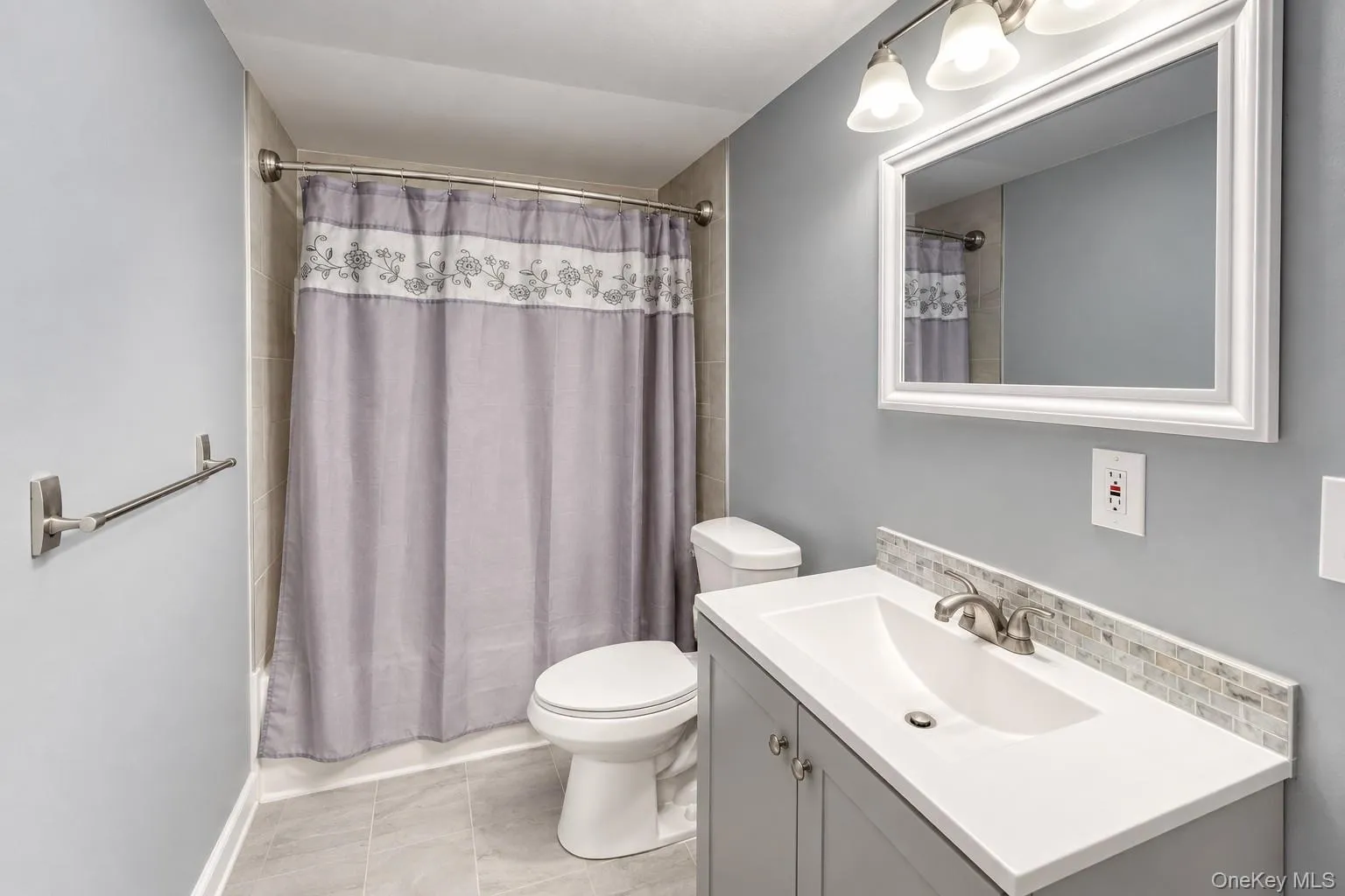 Stylish full bathroom featuring a modern vanity with brushed-nickel fixtures, white framed mirror, and elegant tile backsplash. The space is accented by soft gray walls, coordinating tile flooring, and a decorative shower curtain with floral detailing. Updated lighting and neutral finishes create a bright, contemporary atmosphere that complements the home’s overall design. Stylish full bathroom featuring a modern vanity with brushed-nickel fixtures, white framed mirror, and elegant tile backsplash. The space is accented by soft gray walls, coordinating tile flooring, and a decorative shower curtain with floral detailing. Updated lighting and neutral finishes create a bright, contemporary atmosphere that complements the home’s overall design.