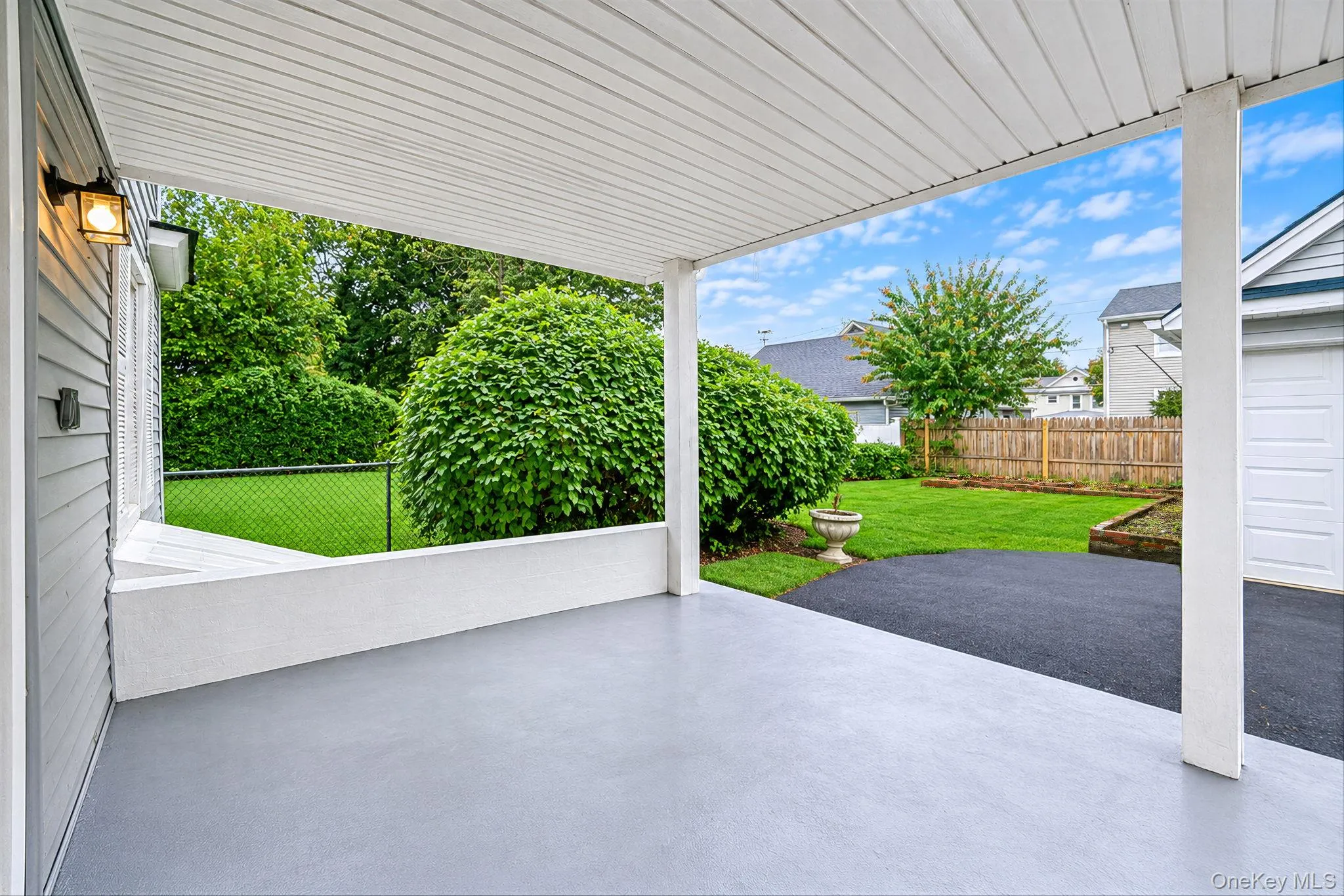 Covered patio featuring a concrete floor, beadboard ceiling, wall-mounted exterior light fixture, and low-profile masonry wall, opening to a fenced backyard with a lawn and asphalt driveway *virtually edited Covered patio featuring a concrete floor, beadboard ceiling, wall-mounted exterior light fixture, and low-profile masonry wall, opening to a fenced backyard with a lawn and asphalt driveway *virtually edited