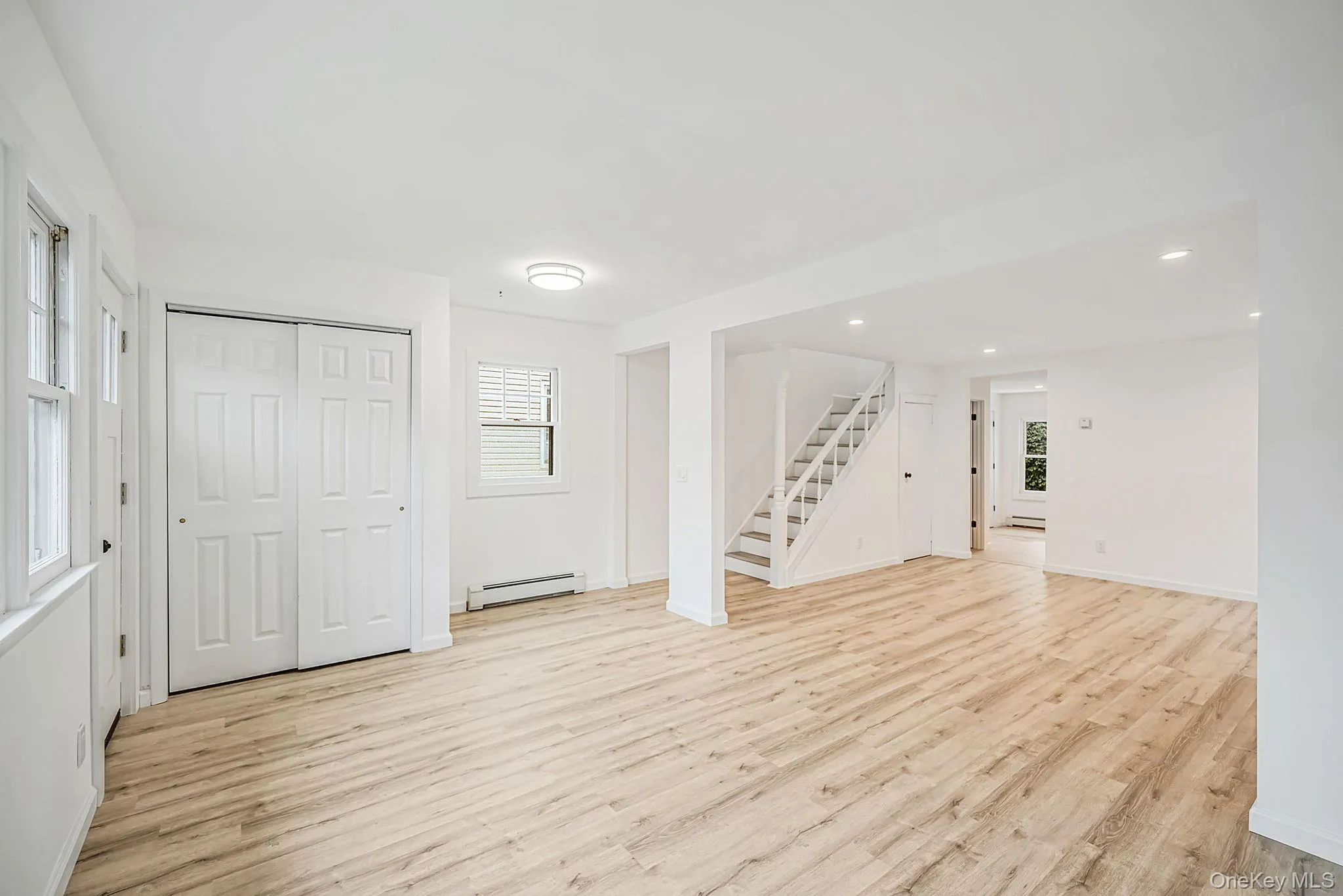 Open-concept interior featuring light-tone wood-finish flooring, recessed lighting, double-panel closet doors, and a staircase with white balusters Open-concept interior featuring light-tone wood-finish flooring, recessed lighting, double-panel closet doors, and a staircase with white balusters