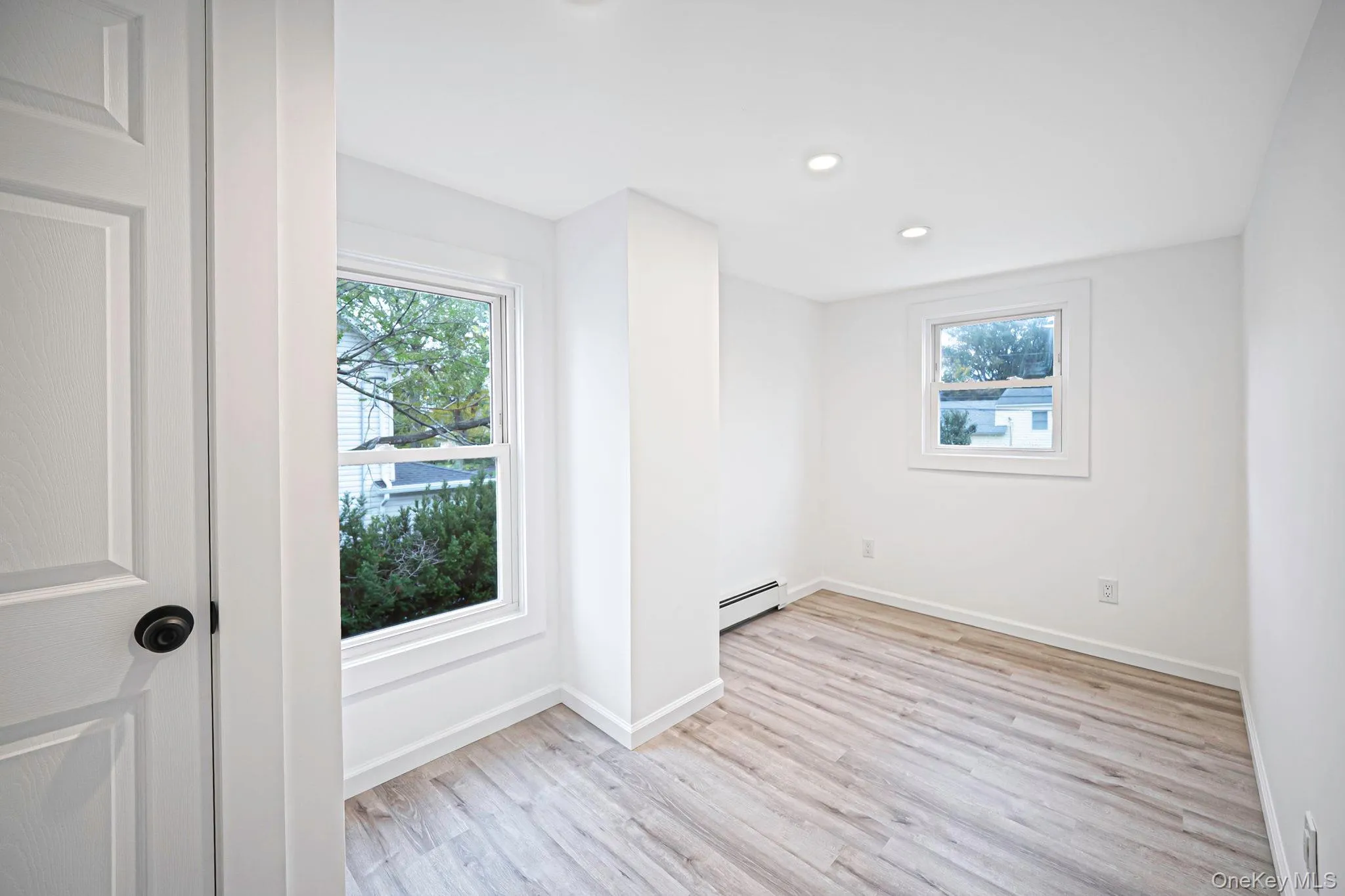 Interior space featuring vinyl wood-finish flooring, recessed lighting, dual windows with white casing, and a paneled interior door with matte black hardware Interior space featuring vinyl wood-finish flooring, recessed lighting, dual windows with white casing, and a paneled interior door with matte black hardware
