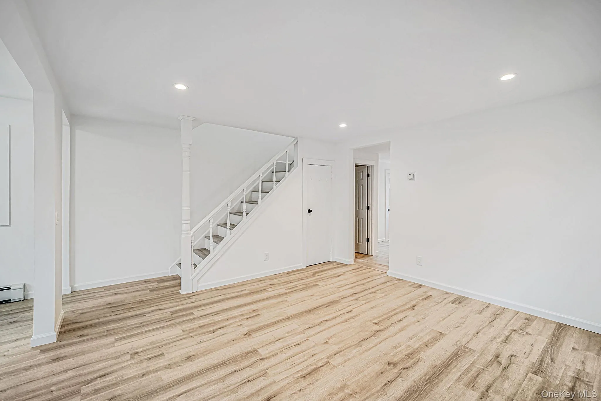 Open-concept interior featuring wood-finish flooring, recessed lighting, and a staircase with white balusters and a decorative post Open-concept interior featuring wood-finish flooring, recessed lighting, and a staircase with white balusters and a decorative post