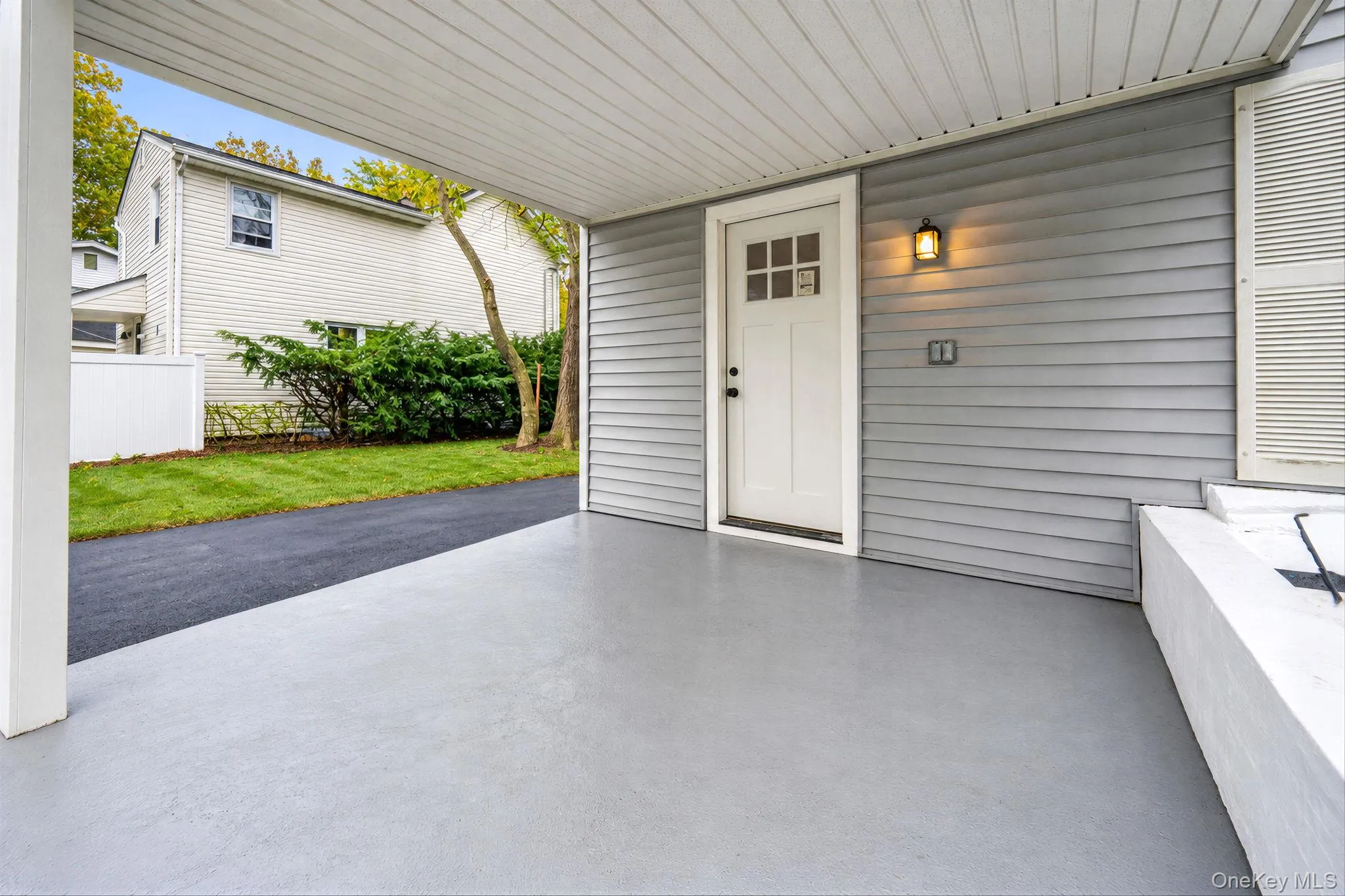 Covered carport featuring gray siding, a painted concrete floor, a six-pane window exterior door, and a wall-mounted light fixture Covered carport featuring gray siding, a painted concrete floor, a six-pane window exterior door, and a wall-mounted light fixture