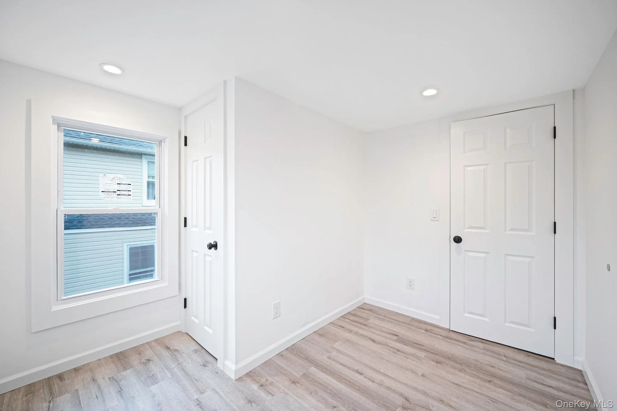 Neutral-toned interior space featuring vinyl wood-finish flooring, recessed lighting, white six-panel doors, and a double-hung window with wide casing Neutral-toned interior space featuring vinyl wood-finish flooring, recessed lighting, white six-panel doors, and a double-hung window with wide casing