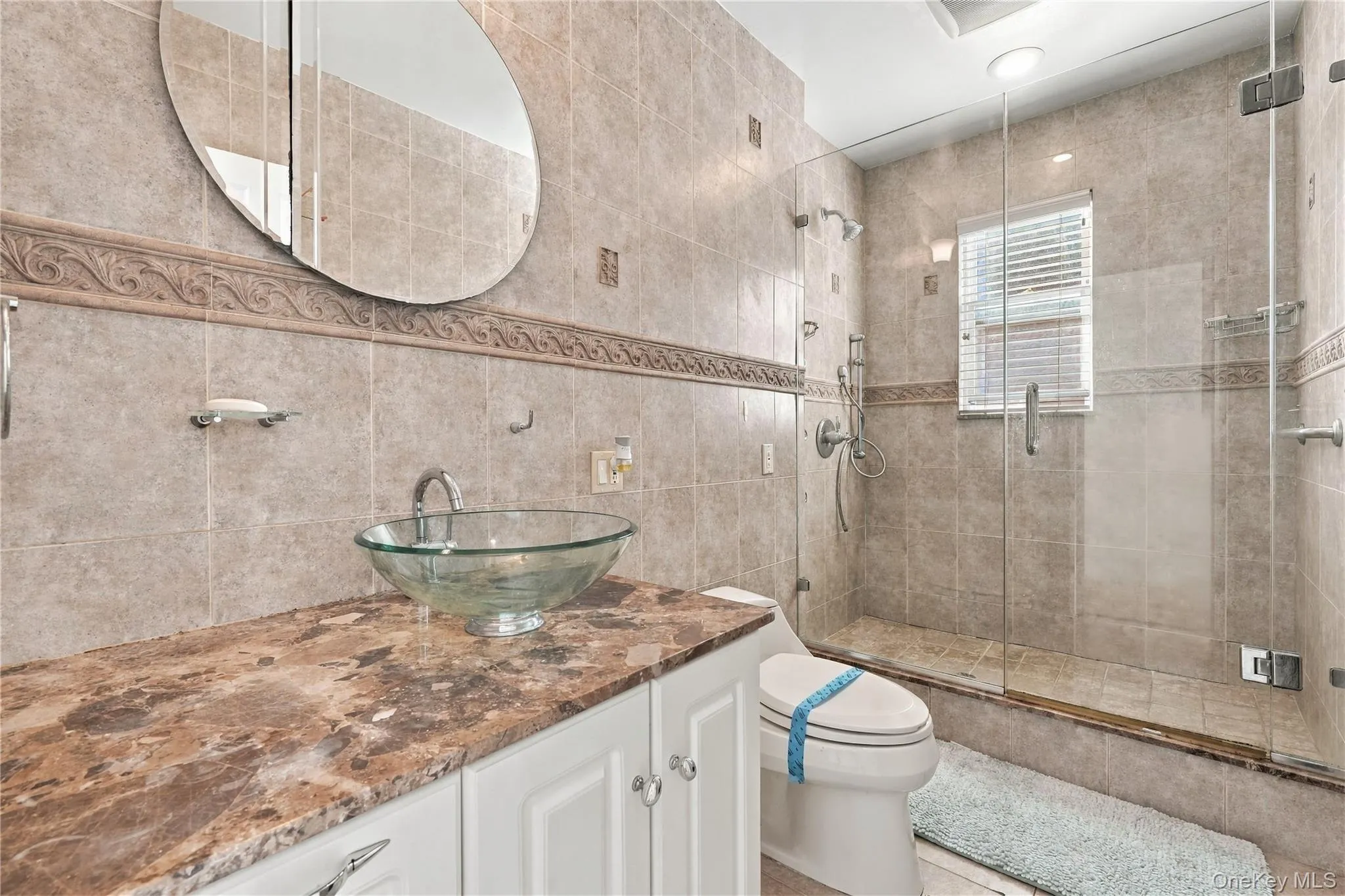 Bathroom featuring a glass vessel sink, stone-finish countertop, white cabinetry, wall-to-wall ceramic tiling with decorative accent trim, and a glass-enclosed shower Bathroom featuring a glass vessel sink, stone-finish countertop, white cabinetry, wall-to-wall ceramic tiling with decorative accent trim, and a glass-enclosed shower