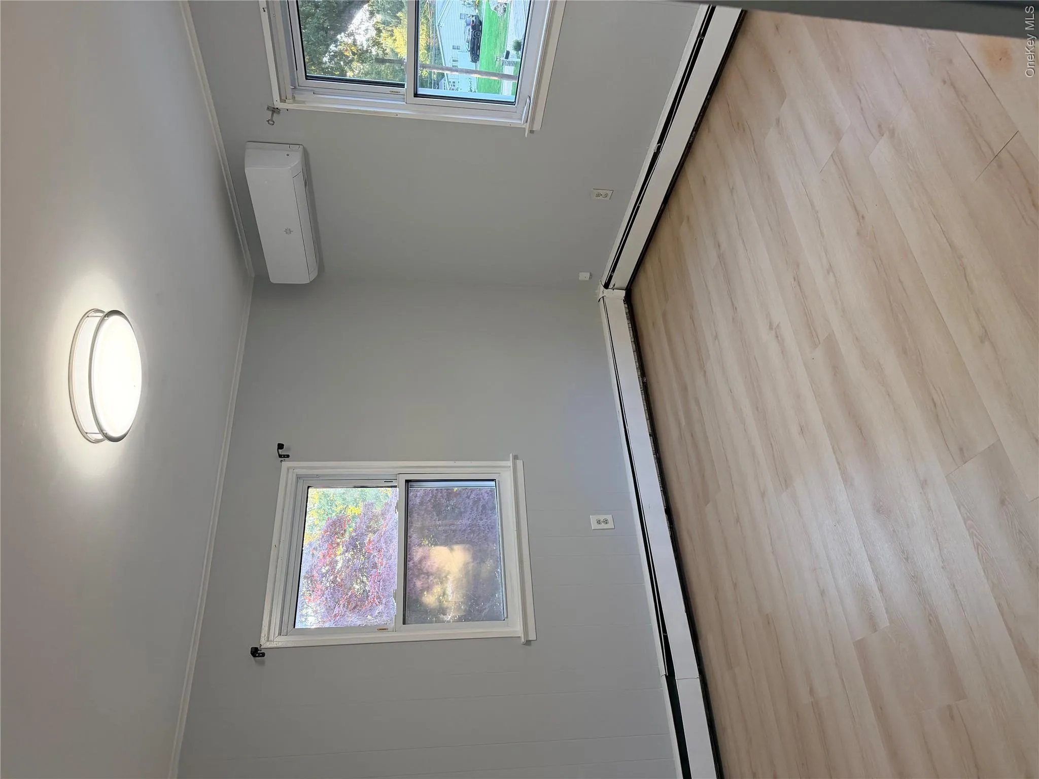 Neutral-toned interior featuring wood-finish flooring, a wall-mounted ductless mini-split system, dual windows, and a flush-mount circular ceiling light fixture Neutral-toned interior featuring wood-finish flooring, a wall-mounted ductless mini-split system, dual windows, and a flush-mount circular ceiling light fixture