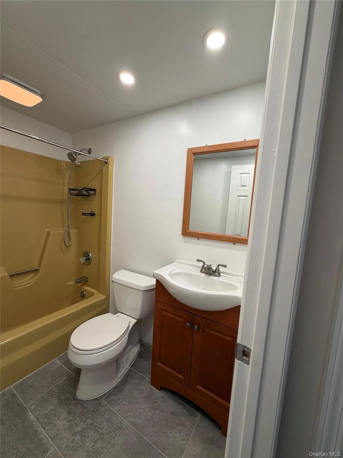 Recessed ceiling lighting, full bathtub with shower, toilet, wood-finish vanity with integrated sink, and gray tile flooring Recessed ceiling lighting, full bathtub with shower, toilet, wood-finish vanity with integrated sink, and gray tile flooring