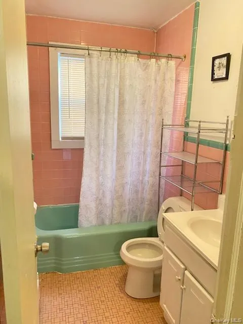 Bathroom featuring a window with blinds, pink wall tiling, a green bathtub, a toilet, and a white vanity with an integrated sink Bathroom featuring a window with blinds, pink wall tiling, a green bathtub, a toilet, and a white vanity with an integrated sink
