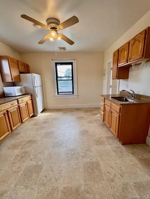 Kitchen with white microwave, dark countertops, brown cabinetry, and a ceiling fan Kitchen with white microwave, dark countertops, brown cabinetry, and a ceiling fan