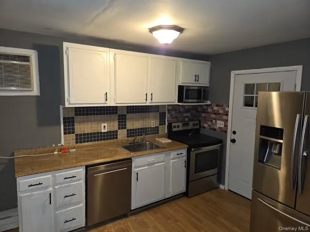 Kitchen with stainless steel appliances, white cabinetry, a wall unit AC, dark wood-style floors, and decorative backsplash Kitchen with stainless steel appliances, white cabinetry, a wall unit AC, dark wood-style floors, and decorative backsplash