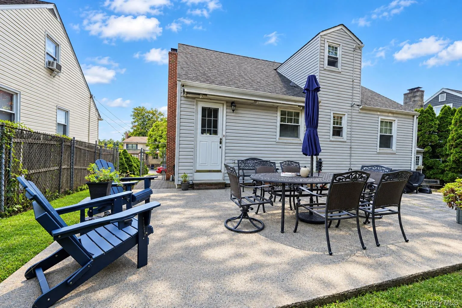 Rear exterior featuring light-toned horizontal siding, a textured concrete patio, a chimney with brick detailing, and an enclosed backyard with a chain-link fence Rear exterior featuring light-toned horizontal siding, a textured concrete patio, a chimney with brick detailing, and an enclosed backyard with a chain-link fence