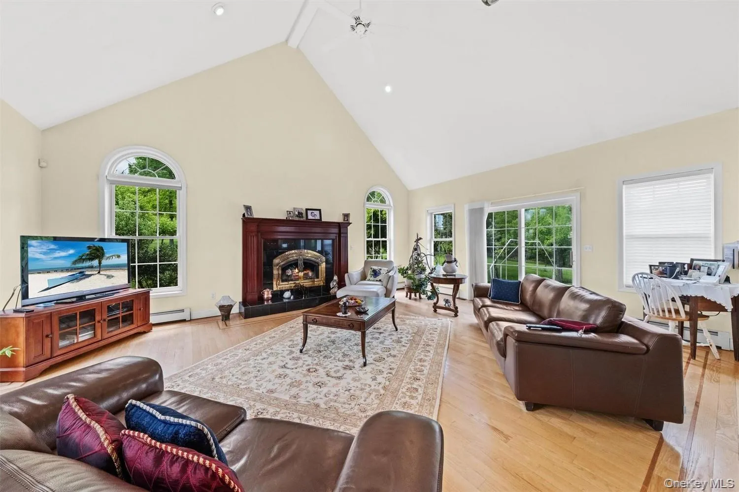 Living room with a high ceiling, light wood-type flooring, a ceiling fan, a premium fireplace, and a baseboard heating unit Living room with a high ceiling, light wood-type flooring, a ceiling fan, a premium fireplace, and a baseboard heating unit