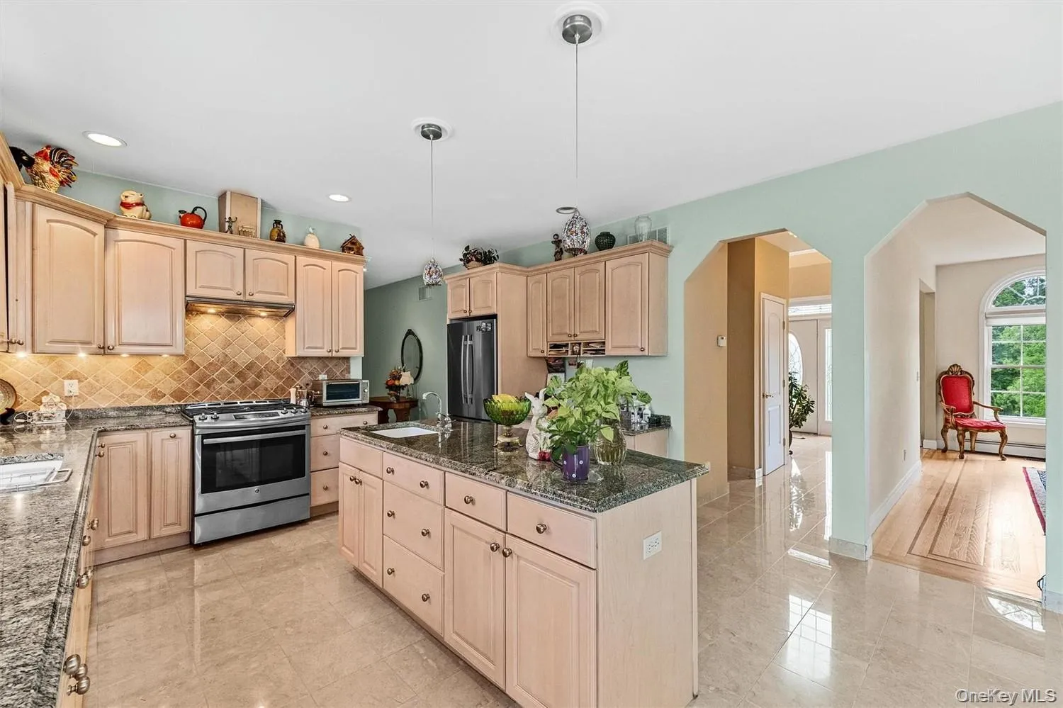 Kitchen featuring arched walkways, light wood finish cabinets, stainless steel appliances, dark stone countertops, and decorative light fixtures Kitchen featuring arched walkways, light wood finish cabinets, stainless steel appliances, dark stone countertops, and decorative light fixtures