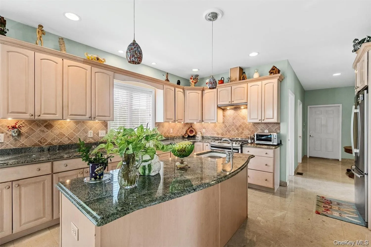 Kitchen with light wood finish cabinetry, a kitchen island with sink, dark stone countertops, hanging light fixtures, and stainless steel appliances Kitchen with light wood finish cabinetry, a kitchen island with sink, dark stone countertops, hanging light fixtures, and stainless steel appliances