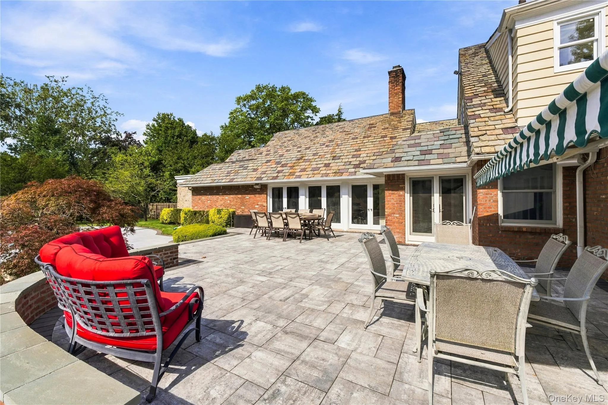 Spacious stone patio featuring a curved brick retaining wall, multi-tonal slate roof tiles, brick exterior siding, and a striped retractable awning Spacious stone patio featuring a curved brick retaining wall, multi-tonal slate roof tiles, brick exterior siding, and a striped retractable awning