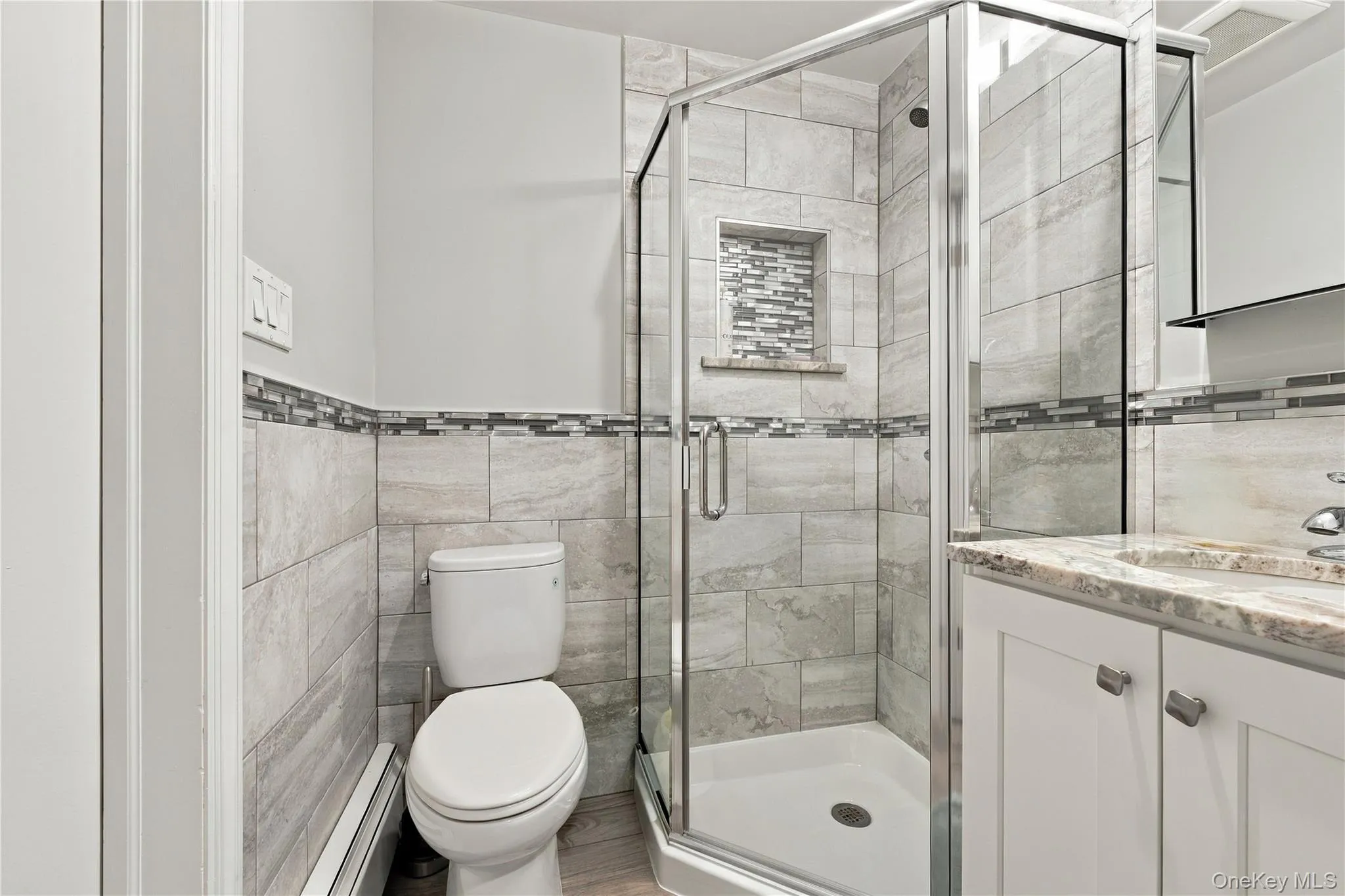 Corner shower featuring a glass enclosure and built-in tiled niche, vanity with stone-finish countertop, and full-height wall tiling with decorative mosaic tile accents Corner shower featuring a glass enclosure and built-in tiled niche, vanity with stone-finish countertop, and full-height wall tiling with decorative mosaic tile accents