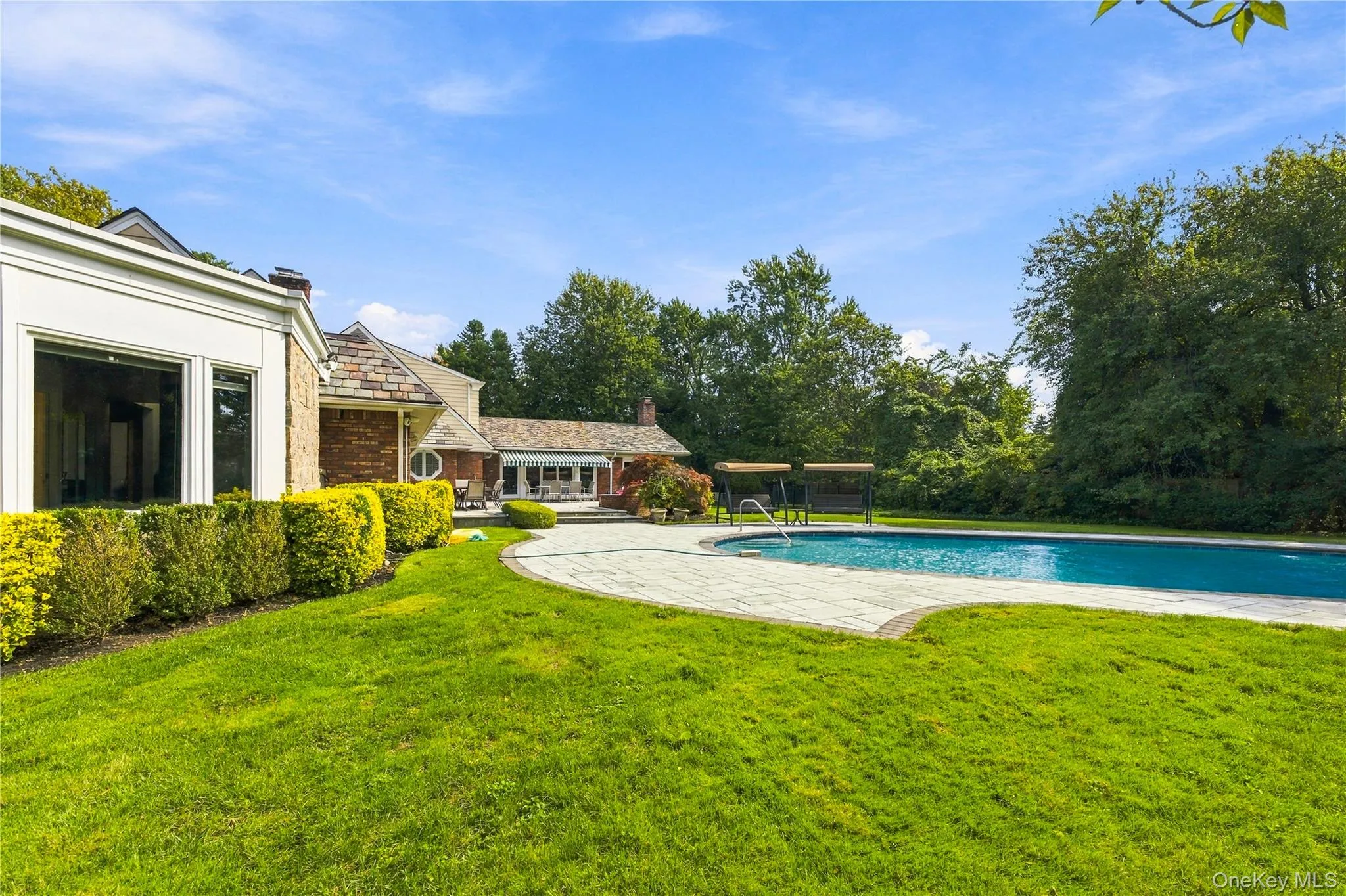 Expansive rear grounds featuring a curved stone pool deck, in-ground swimming pool, manicured lawn, and mature perimeter trees Expansive rear grounds featuring a curved stone pool deck, in-ground swimming pool, manicured lawn, and mature perimeter trees