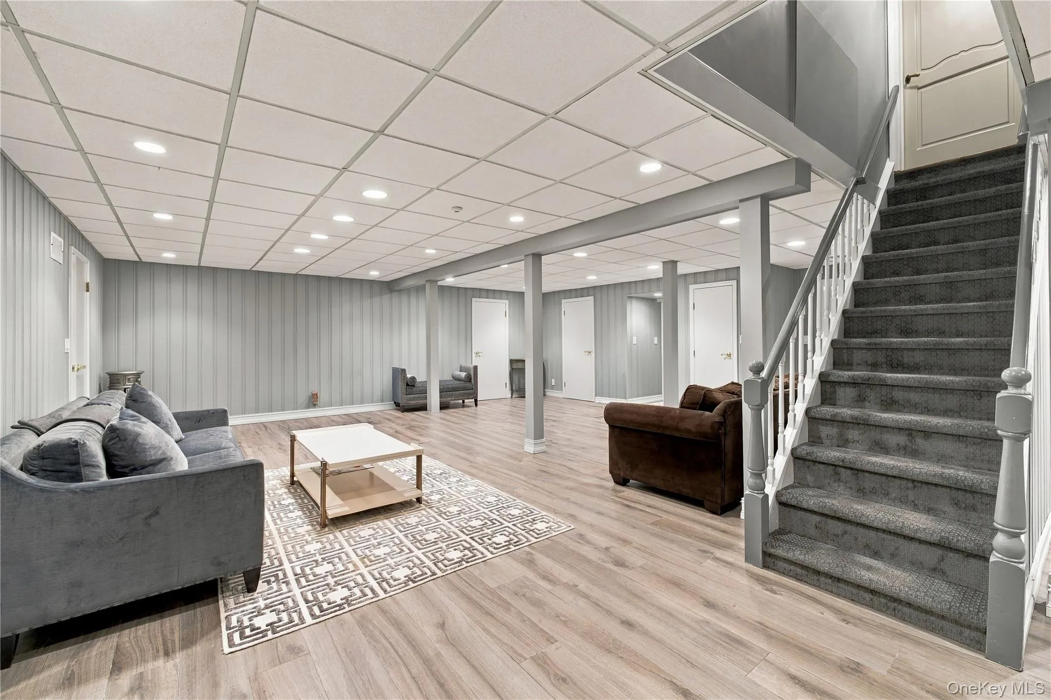 Finished space featuring wood-finish flooring, a drop ceiling with recessed lighting, vertical paneled wall treatments, and a carpeted staircase with painted wood railings Finished space featuring wood-finish flooring, a drop ceiling with recessed lighting, vertical paneled wall treatments, and a carpeted staircase with painted wood railings