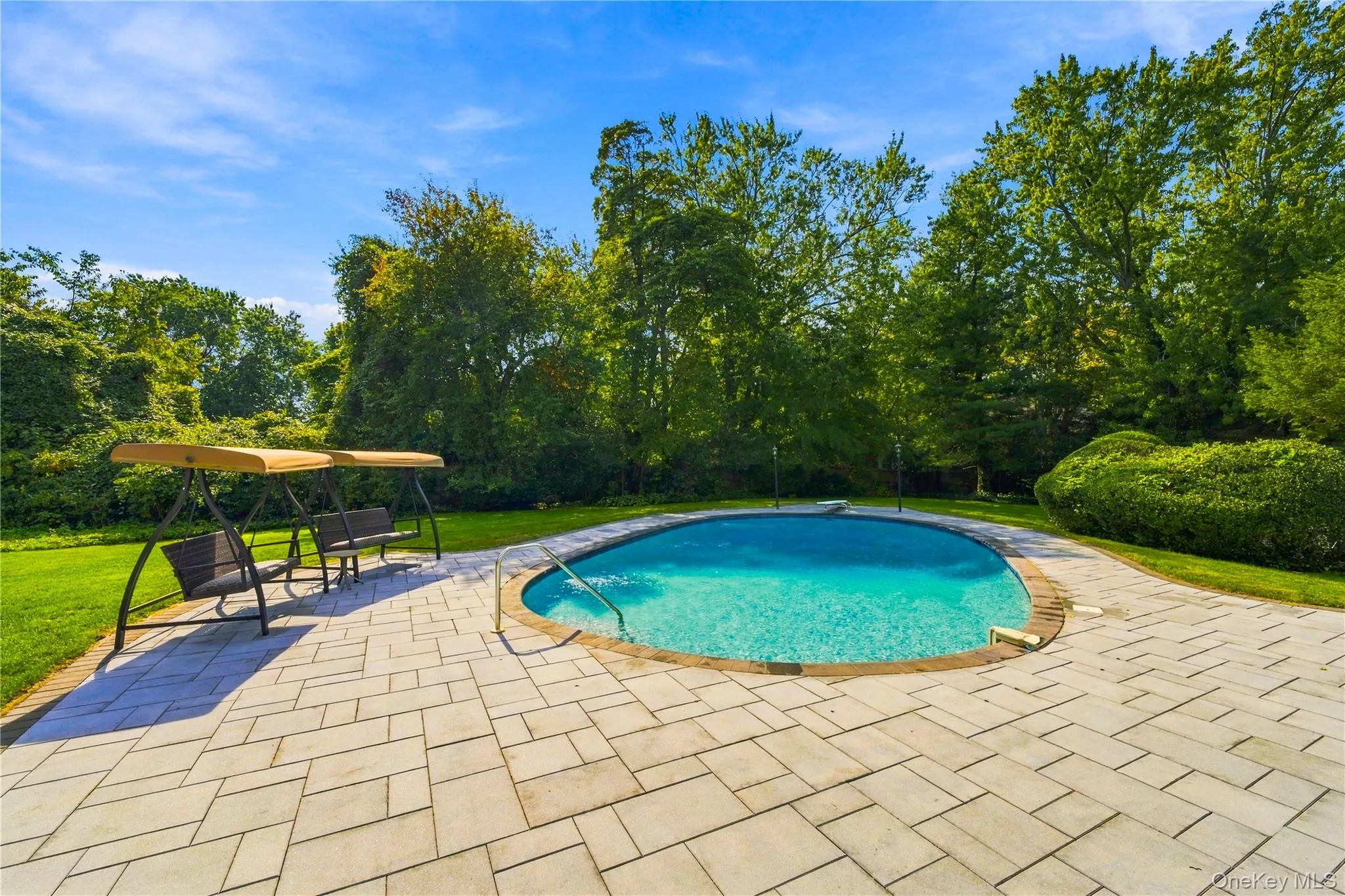 Inground swimming pool with metal ladder, surrounded by an expansive stone paver patio, set within a level lawn bordered by mature trees and shrubs Inground swimming pool with metal ladder, surrounded by an expansive stone paver patio, set within a level lawn bordered by mature trees and shrubs