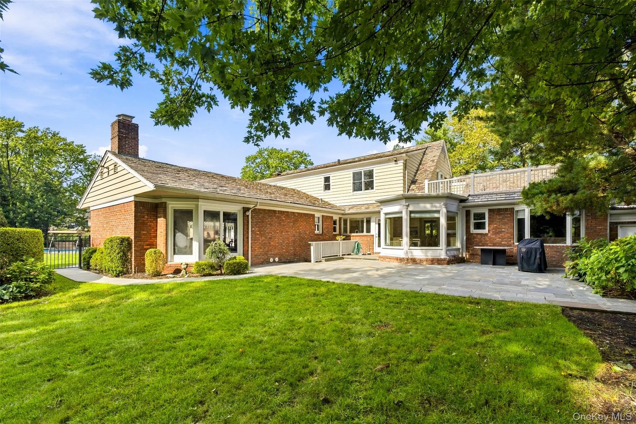 Rear elevation featuring a brick and light-toned horizontal siding exterior, expansive stone patio, masonry chimney, and upper-level railed terrace Rear elevation featuring a brick and light-toned horizontal siding exterior, expansive stone patio, masonry chimney, and upper-level railed terrace