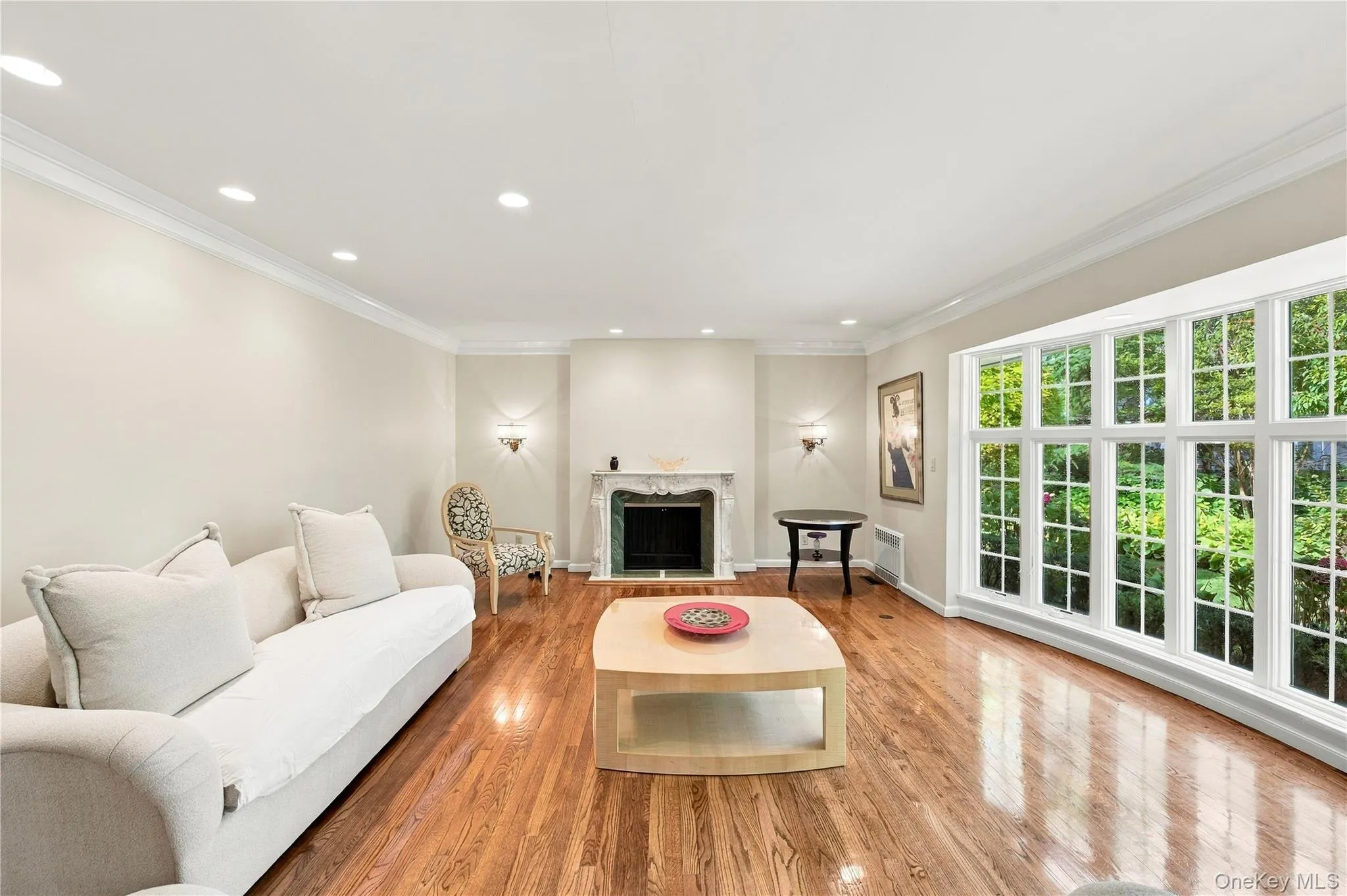 Living area featuring hardwood flooring, a decorative stone-surround fireplace, crown molding, recessed lighting, and expansive multi-pane windows Living area featuring hardwood flooring, a decorative stone-surround fireplace, crown molding, recessed lighting, and expansive multi-pane windows