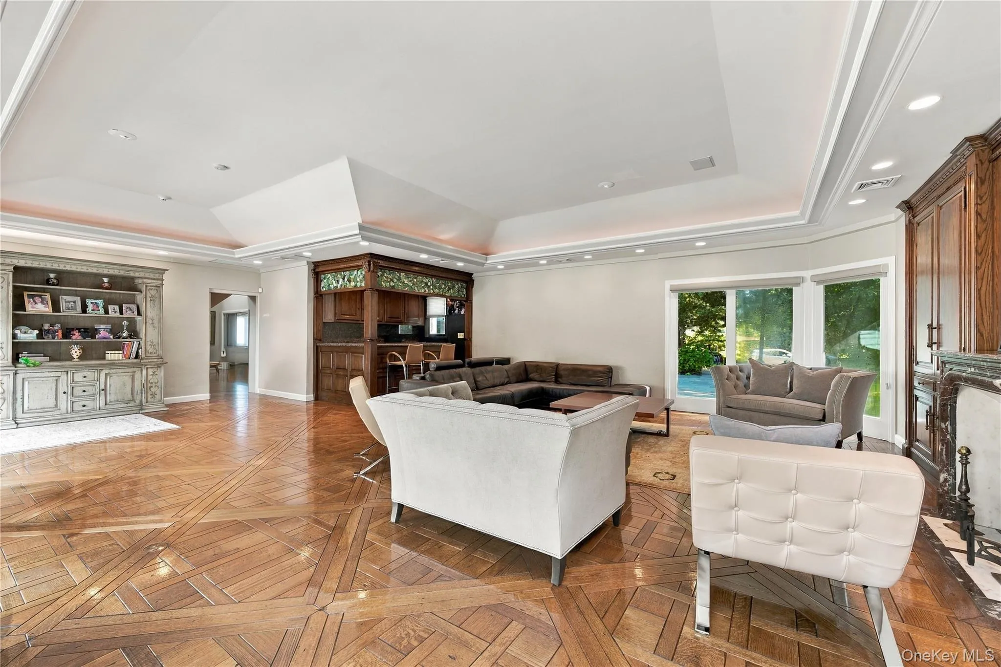 Expansive room featuring intricate parquet wood-finish flooring, a tiered tray ceiling with recessed lighting, custom built-in cabinetry, a wet bar with integrated shelving, and a stone-surround fireplace Expansive room featuring intricate parquet wood-finish flooring, a tiered tray ceiling with recessed lighting, custom built-in cabinetry, a wet bar with integrated shelving, and a stone-surround fireplace