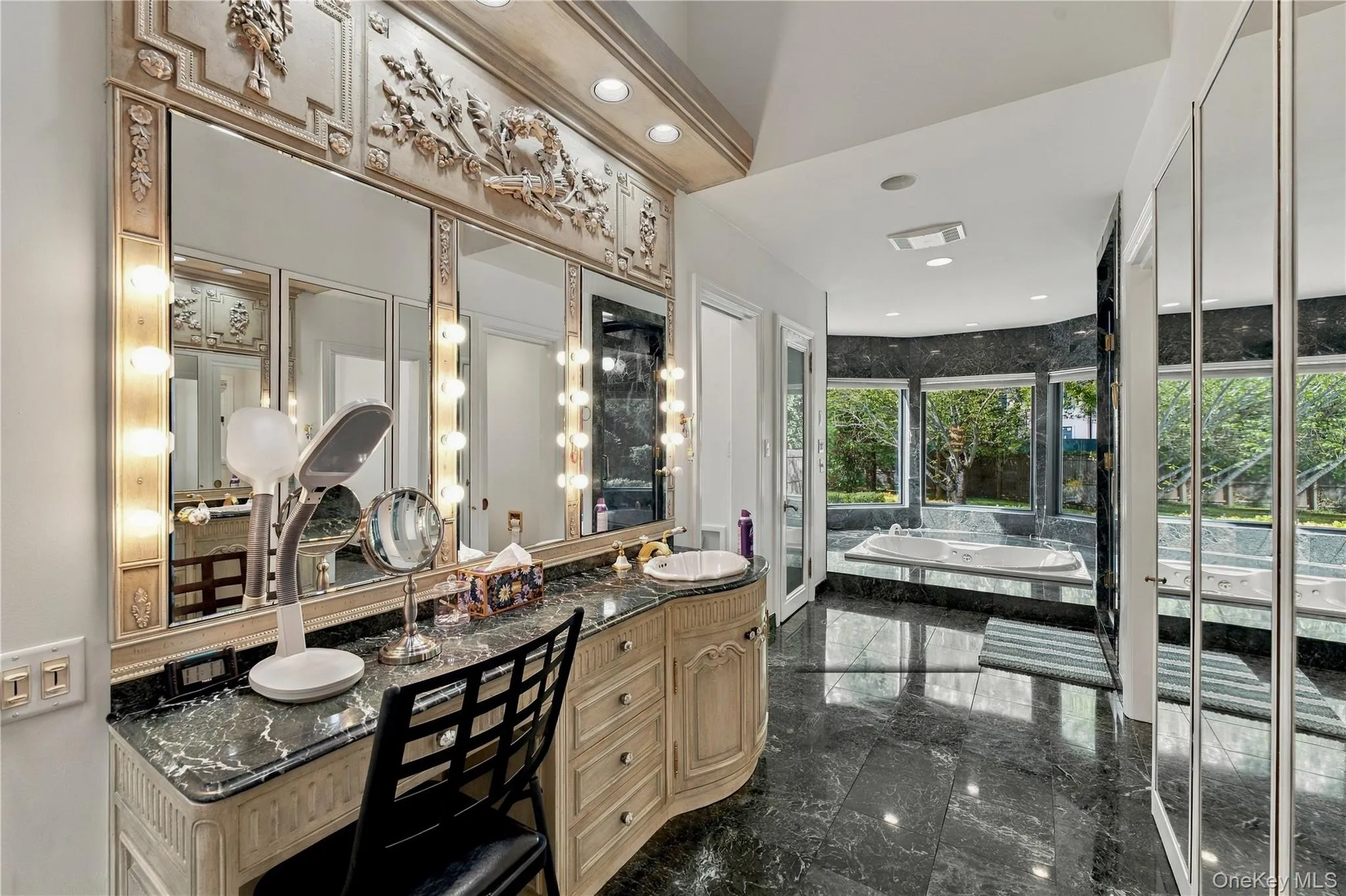 Custom-built vanity with stone countertop and integrated lighting, featuring an ornate architectural mirror frame, polished stone flooring, and a jetted soaking tub set within a multi-paneled window surround Custom-built vanity with stone countertop and integrated lighting, featuring an ornate architectural mirror frame, polished stone flooring, and a jetted soaking tub set within a multi-paneled window surround