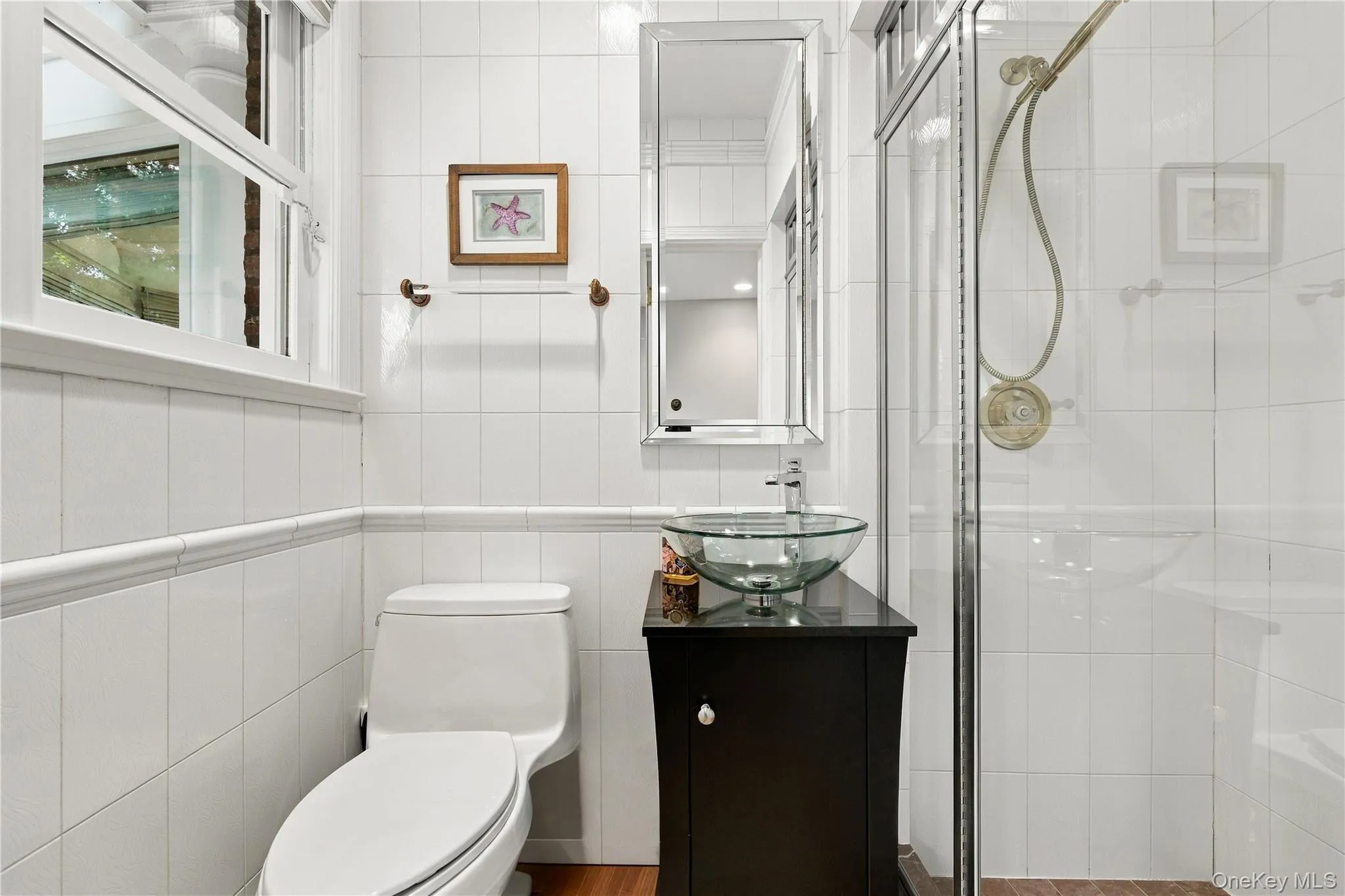 Bathroom featuring white wall tiling with chair rail molding, a dark wood-finish vanity with a glass vessel sink, a chrome faucet, and a glass-enclosed shower with a handheld showerhead Bathroom featuring white wall tiling with chair rail molding, a dark wood-finish vanity with a glass vessel sink, a chrome faucet, and a glass-enclosed shower with a handheld showerhead