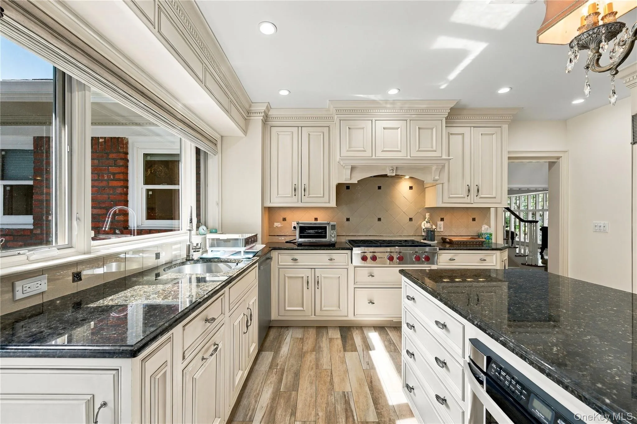 Kitchen featuring cream-toned cabinetry with crown molding, dark stone countertops, wood-finish flooring, a stainless steel range with a custom hood enclosure, and recessed lighting Kitchen featuring cream-toned cabinetry with crown molding, dark stone countertops, wood-finish flooring, a stainless steel range with a custom hood enclosure, and recessed lighting