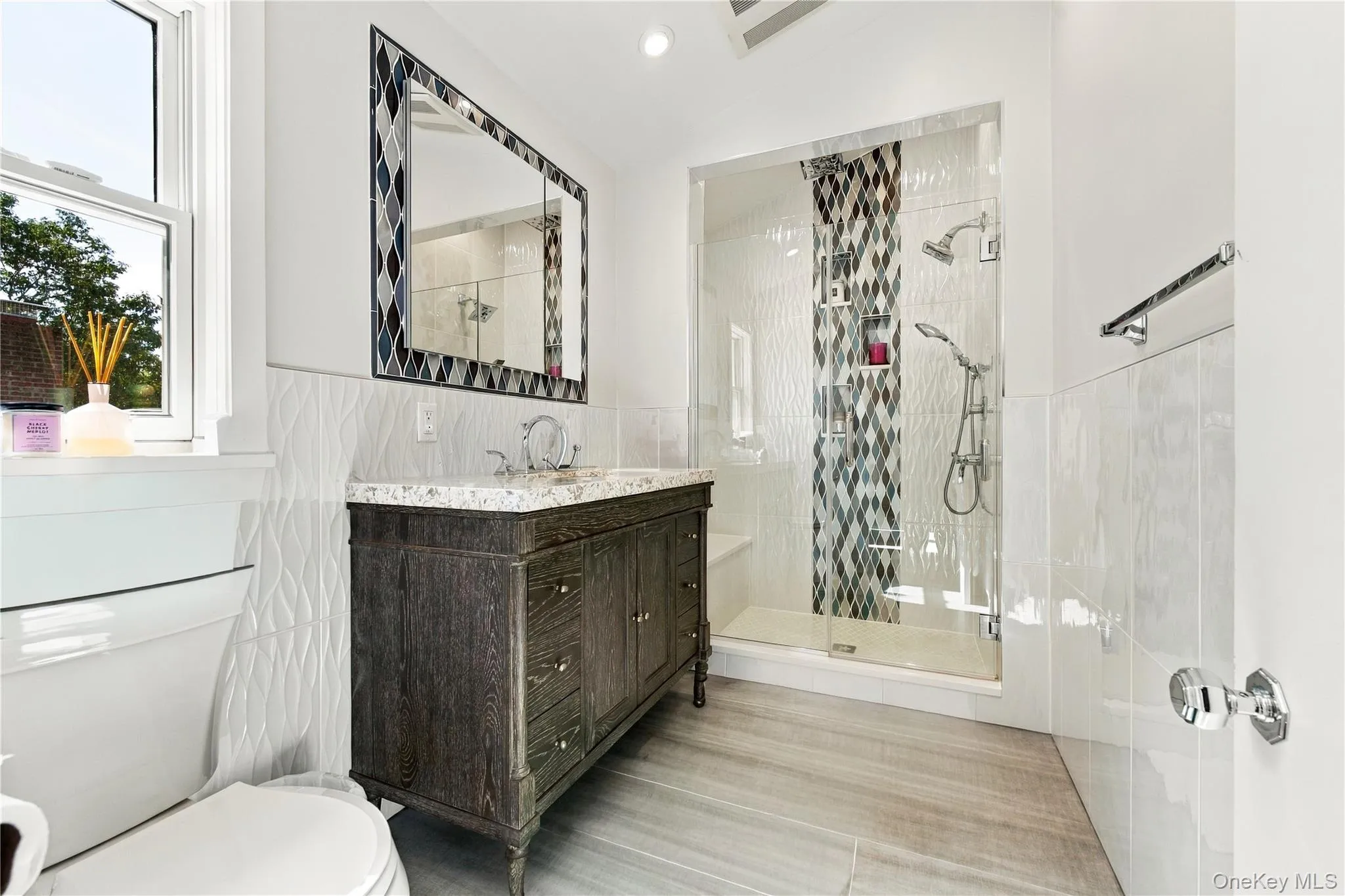 Dark wood-finish vanity with a stone countertop, glass-enclosed shower with mosaic tile inlay, decorative mosaic-framed mirror, and textured wall tile Dark wood-finish vanity with a stone countertop, glass-enclosed shower with mosaic tile inlay, decorative mosaic-framed mirror, and textured wall tile