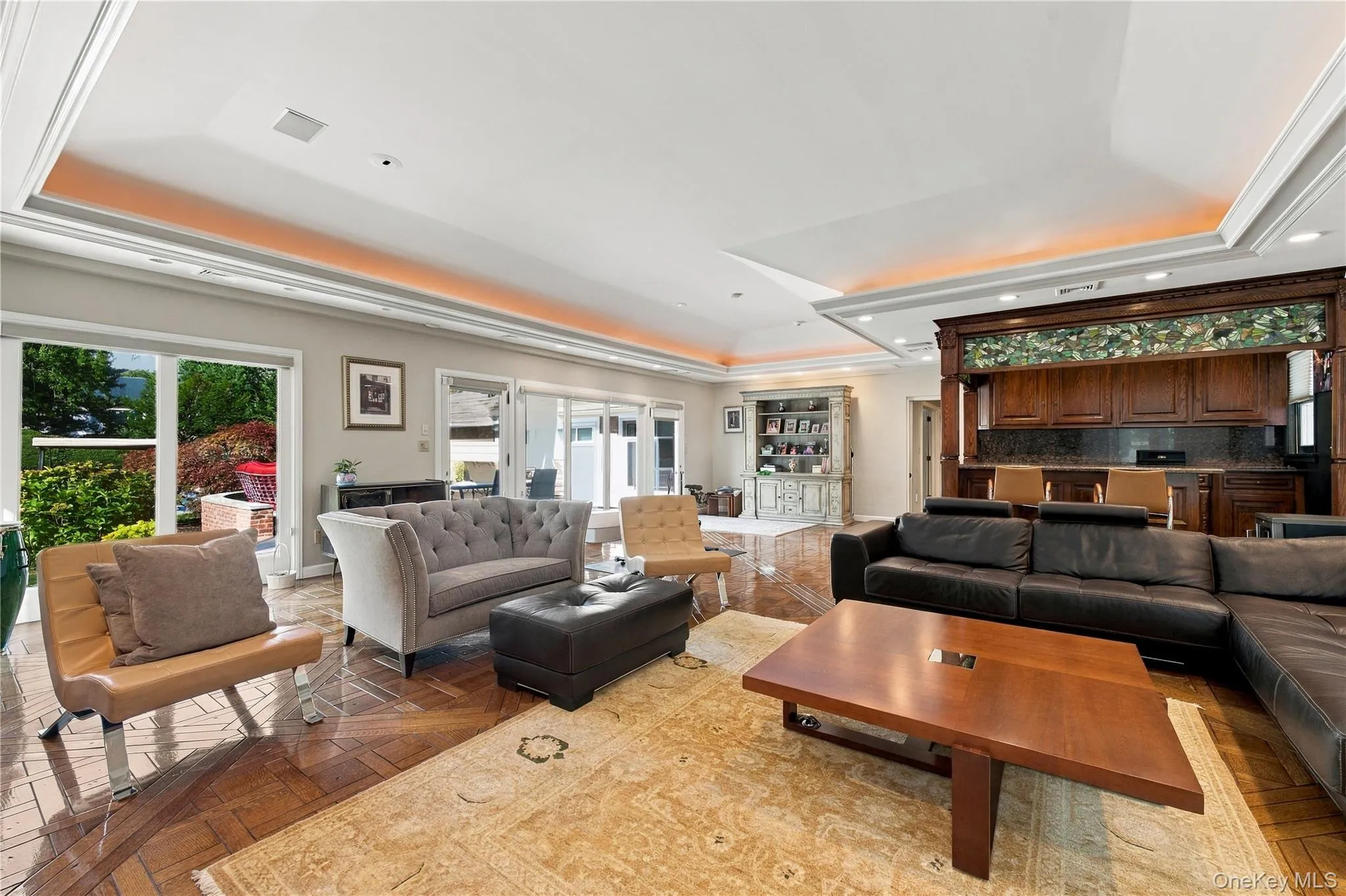 Expansive interior space featuring a multi-tiered tray ceiling with cove lighting, intricate parquet wood flooring, and a custom built-in wooden bar with decorative glass detailing Expansive interior space featuring a multi-tiered tray ceiling with cove lighting, intricate parquet wood flooring, and a custom built-in wooden bar with decorative glass detailing